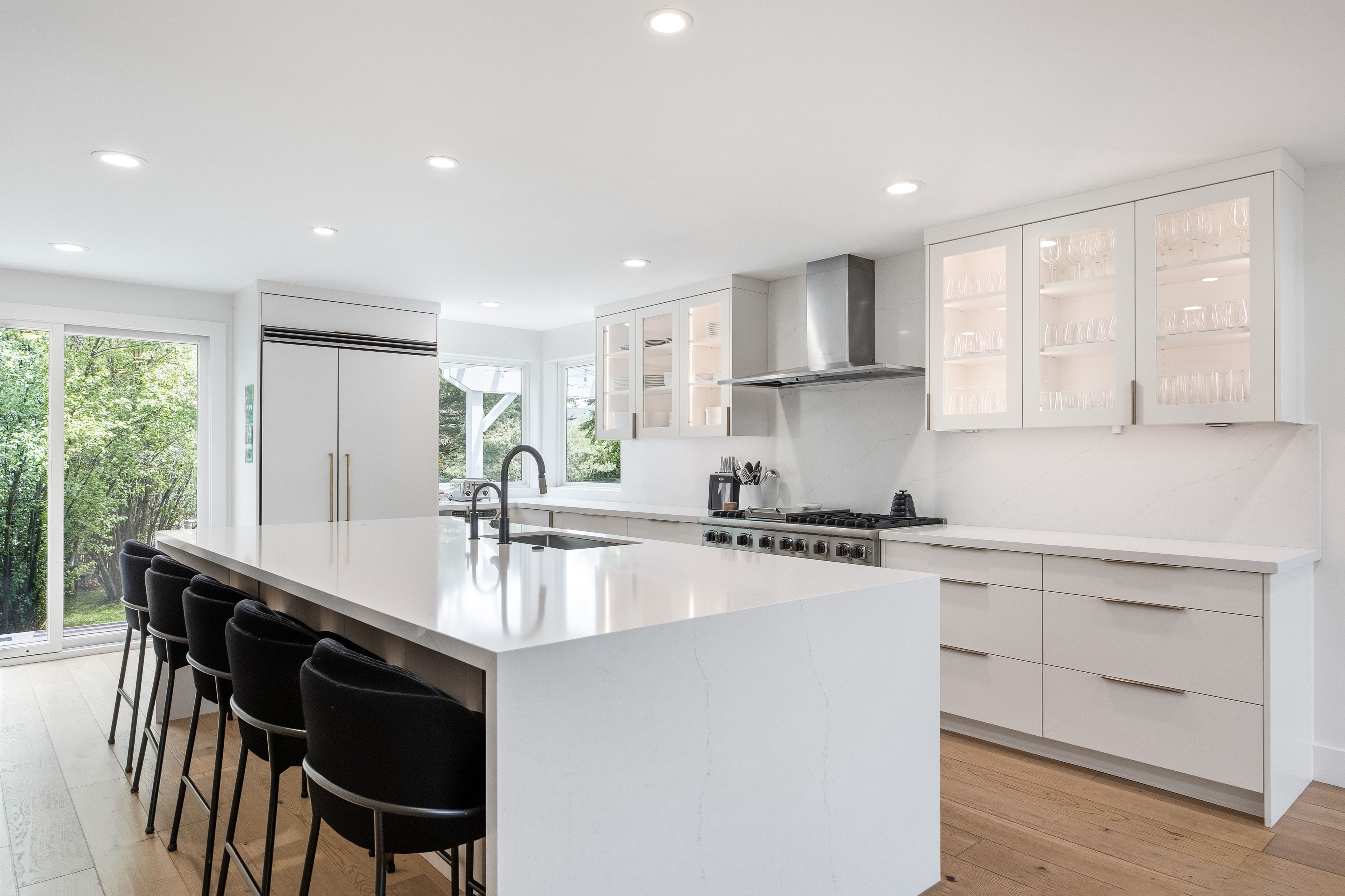 Modern kitchen featuring a large white island with integrated sink, stainless steel appliances, white cabinetry with glass-front uppers, and wood-finish flooring