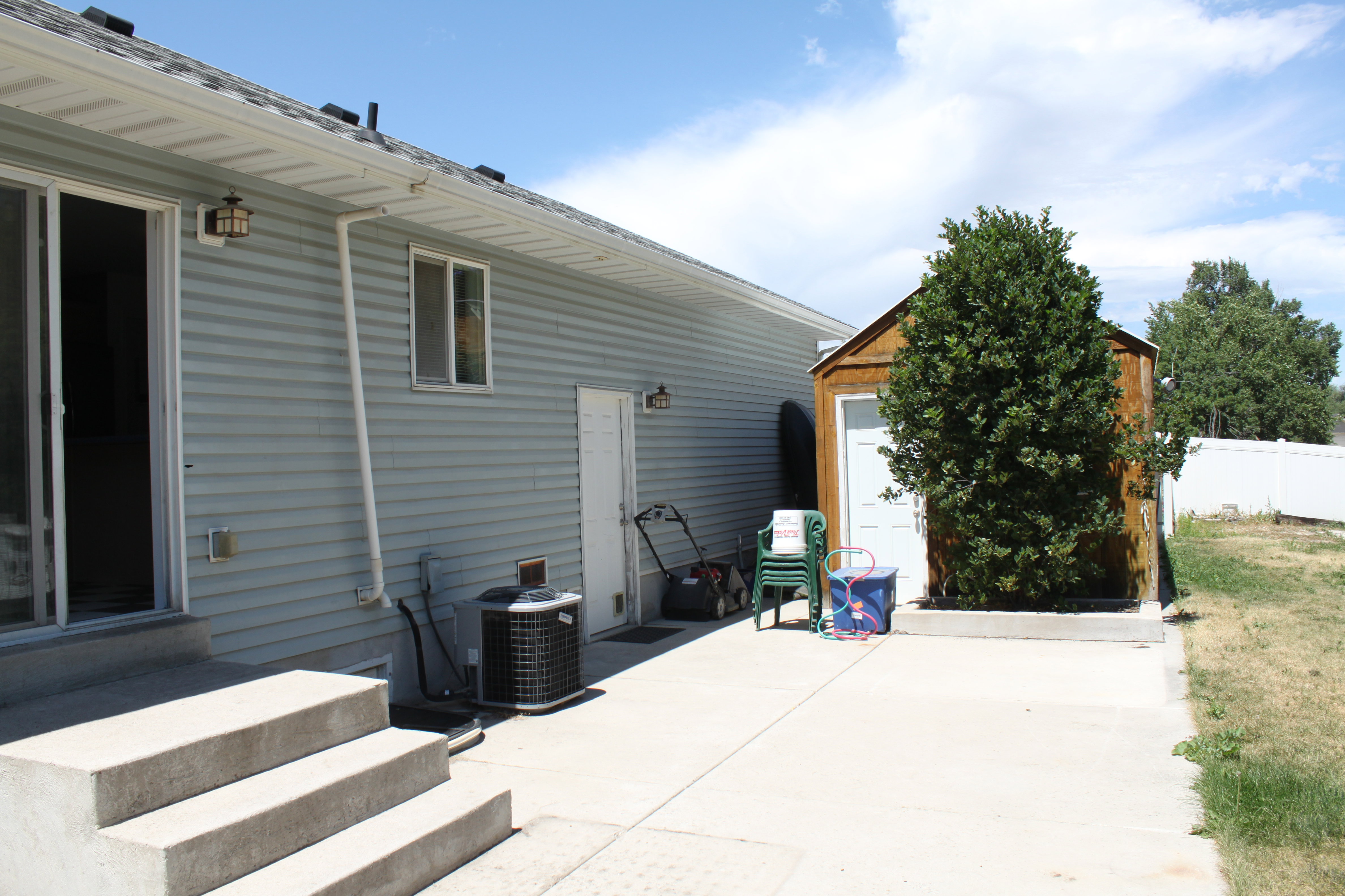 Rear exterior featuring light blue siding, a sliding glass door, a secondary exterior door, and a built-in storage shed with an evergreen shrub
