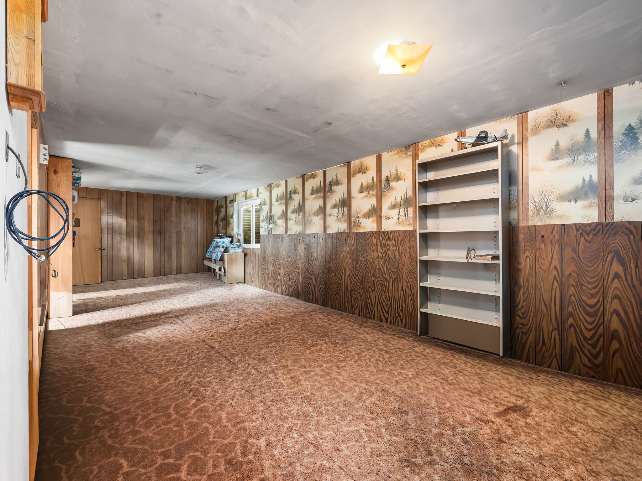 Spacious room featuring patterned carpeting, partial wood paneling, a small window with horizontal blinds, a ceiling-mounted light fixture, and a built-in shelving unit
