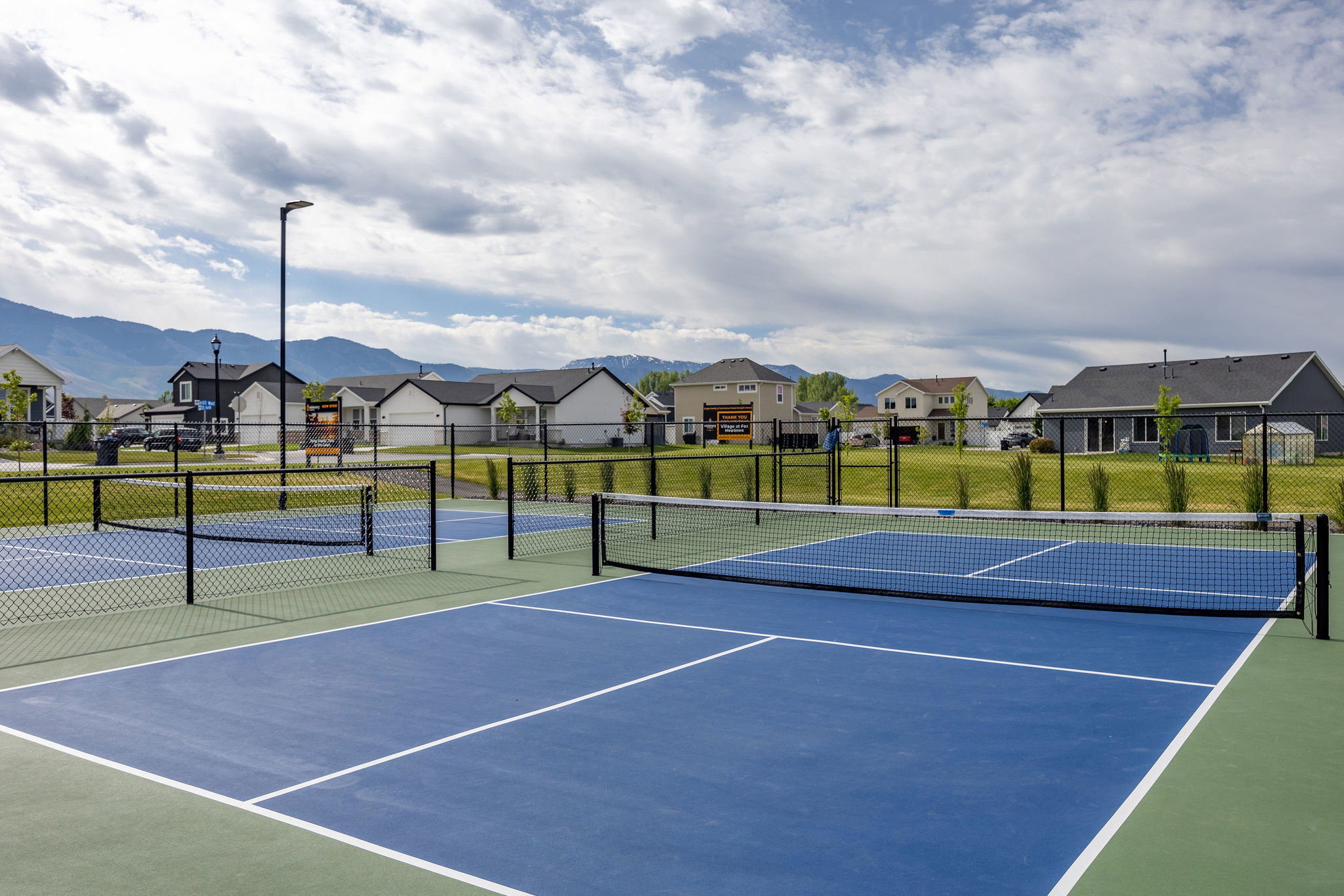 Pickleball court featuring a blue playing surface with white lines, green perimeter, black chain-link fencing, and a permanent net system