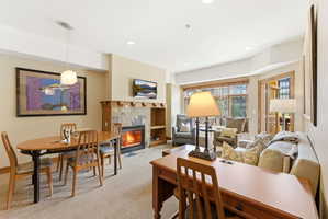 Open-concept living area featuring a stone-clad fireplace with a natural wood mantel and built-in shelving, a dedicated dining space with pendant lighting, recessed ceiling lights, and large windows