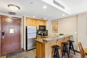 Kitchenette featuring stone-look flooring, light wood cabinetry, stainless steel appliances, a breakfast bar with stone-look countertop, and recessed lighting