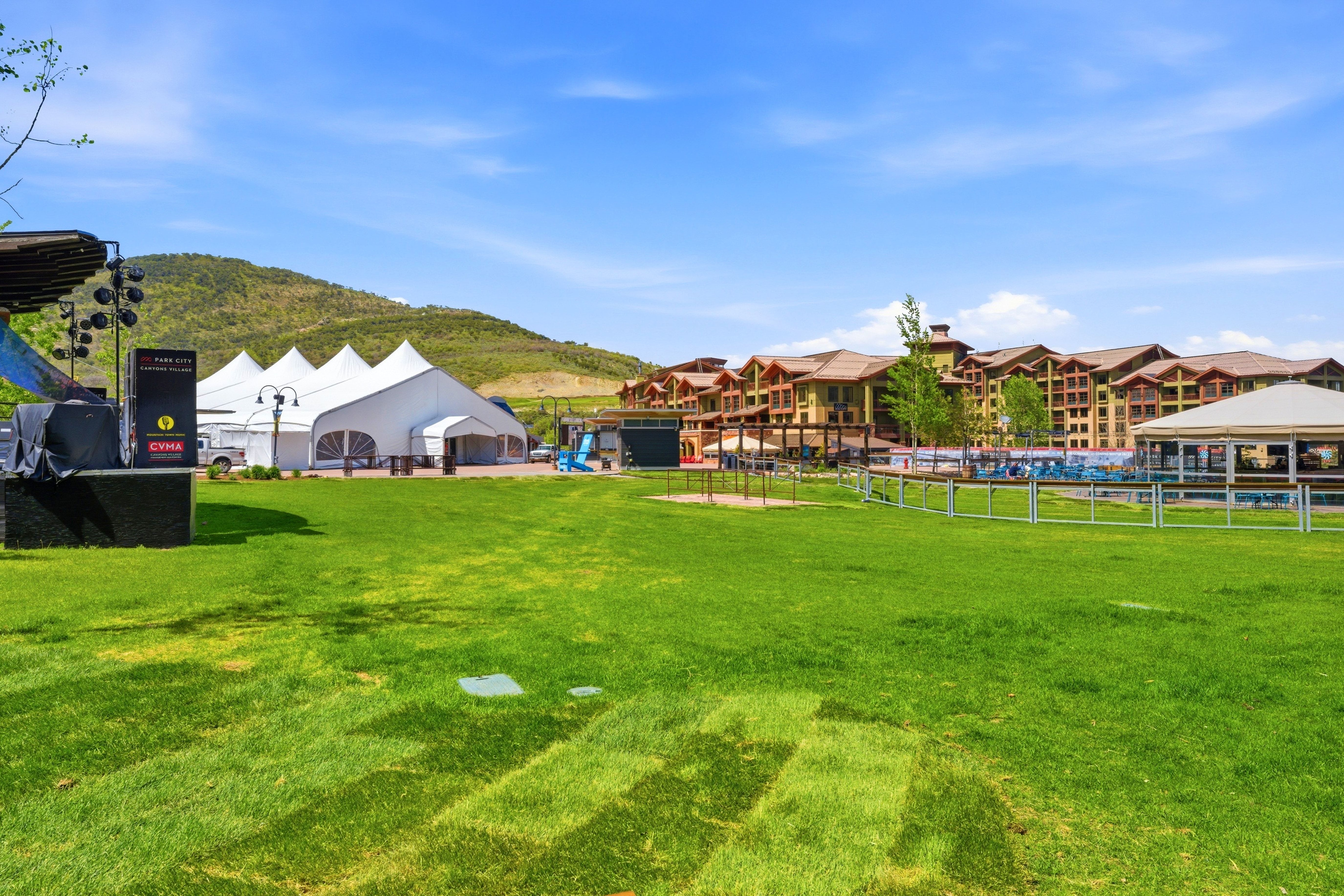 Expansive green lawn surrounded by a white picket fence, with a large event tent and multi-story buildings in the background