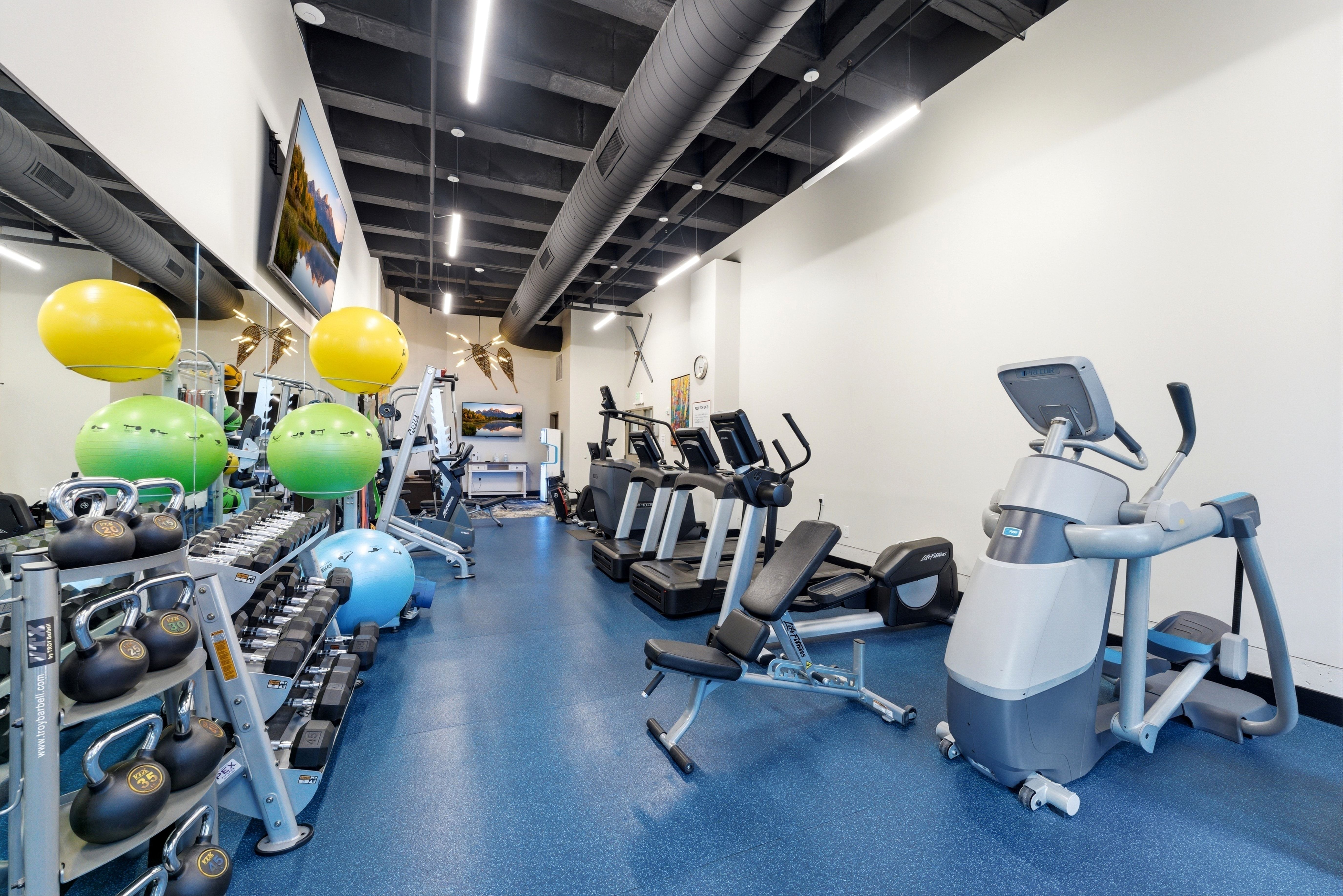 Fitness center featuring blue rubberized flooring, exposed ductwork, modern track lighting, and a mirrored wall