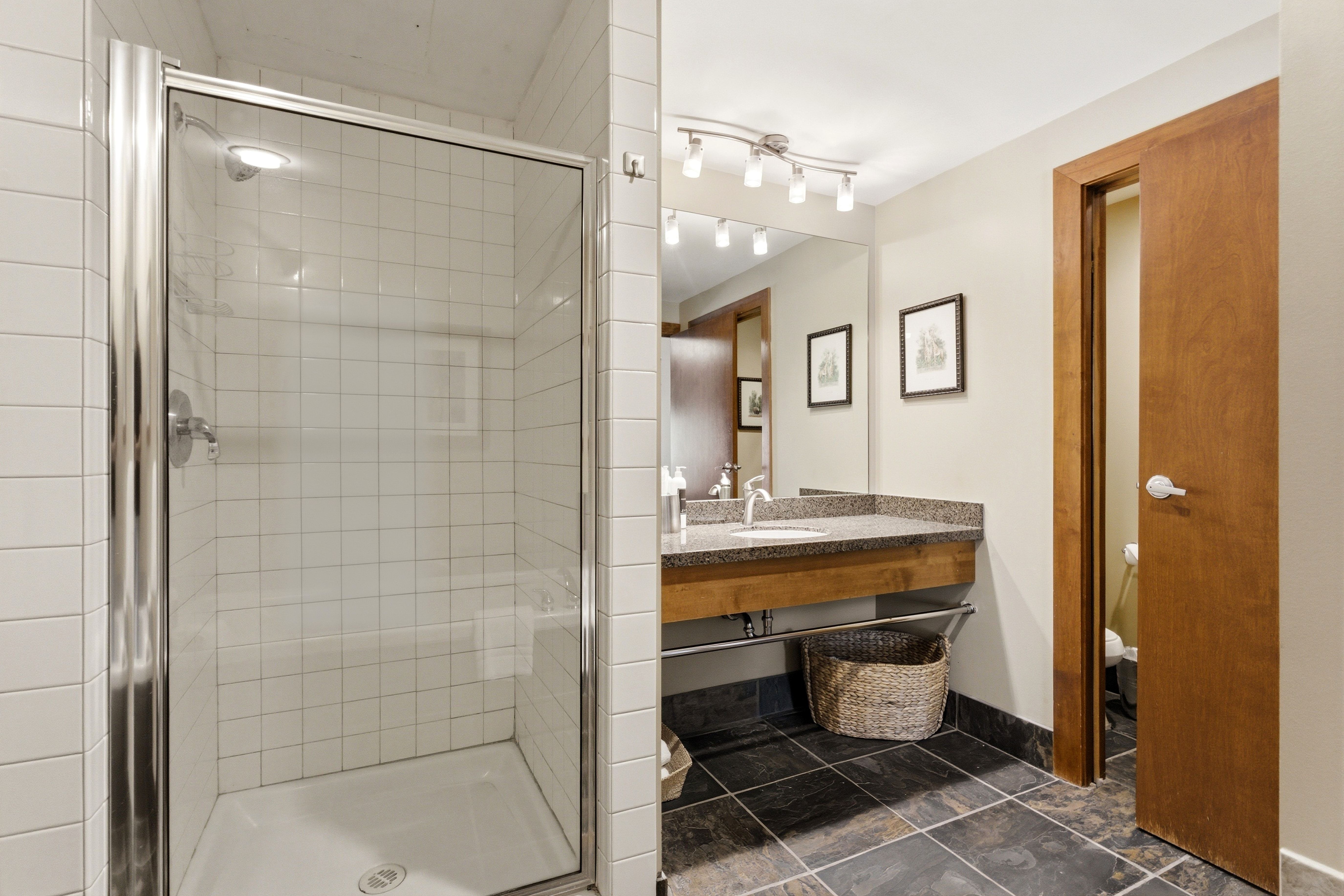 Spacious bathroom featuring a glass-enclosed shower with white subway tile, a vanity with a stone-finish countertop, wood-finish cabinetry, and dark tile flooring