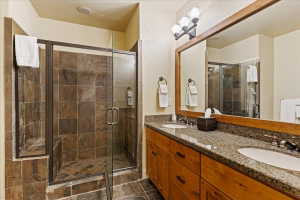 Bathroom featuring a walk-in shower with glass enclosure and stone-finish tile, dual vanity with wood cabinetry, and a large framed mirror with three-bulb vanity lighting