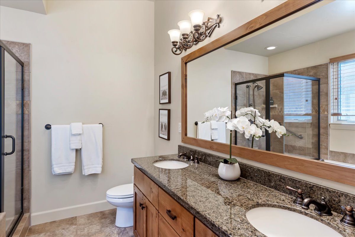 Spacious bathroom featuring a dual vanity with granite-style countertop, wood-finish cabinetry, a large framed mirror, and a glass-enclosed shower with tiled walls
