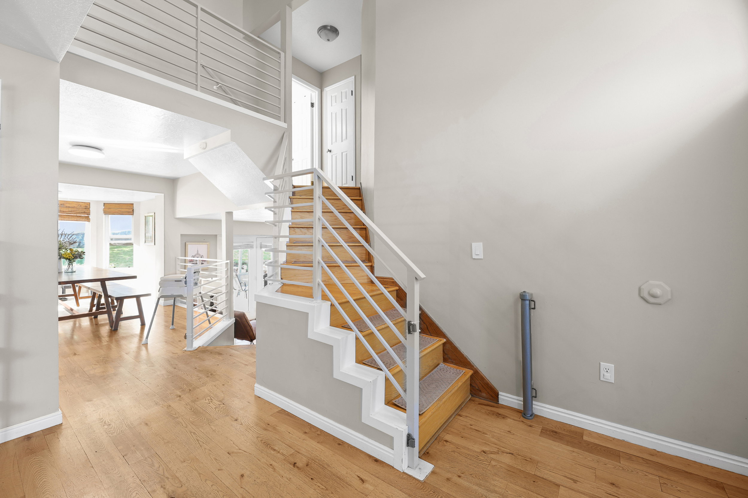 Open-concept interior featuring light wood-finish flooring, a contemporary staircase with horizontal metal railings, and a bay window in the background