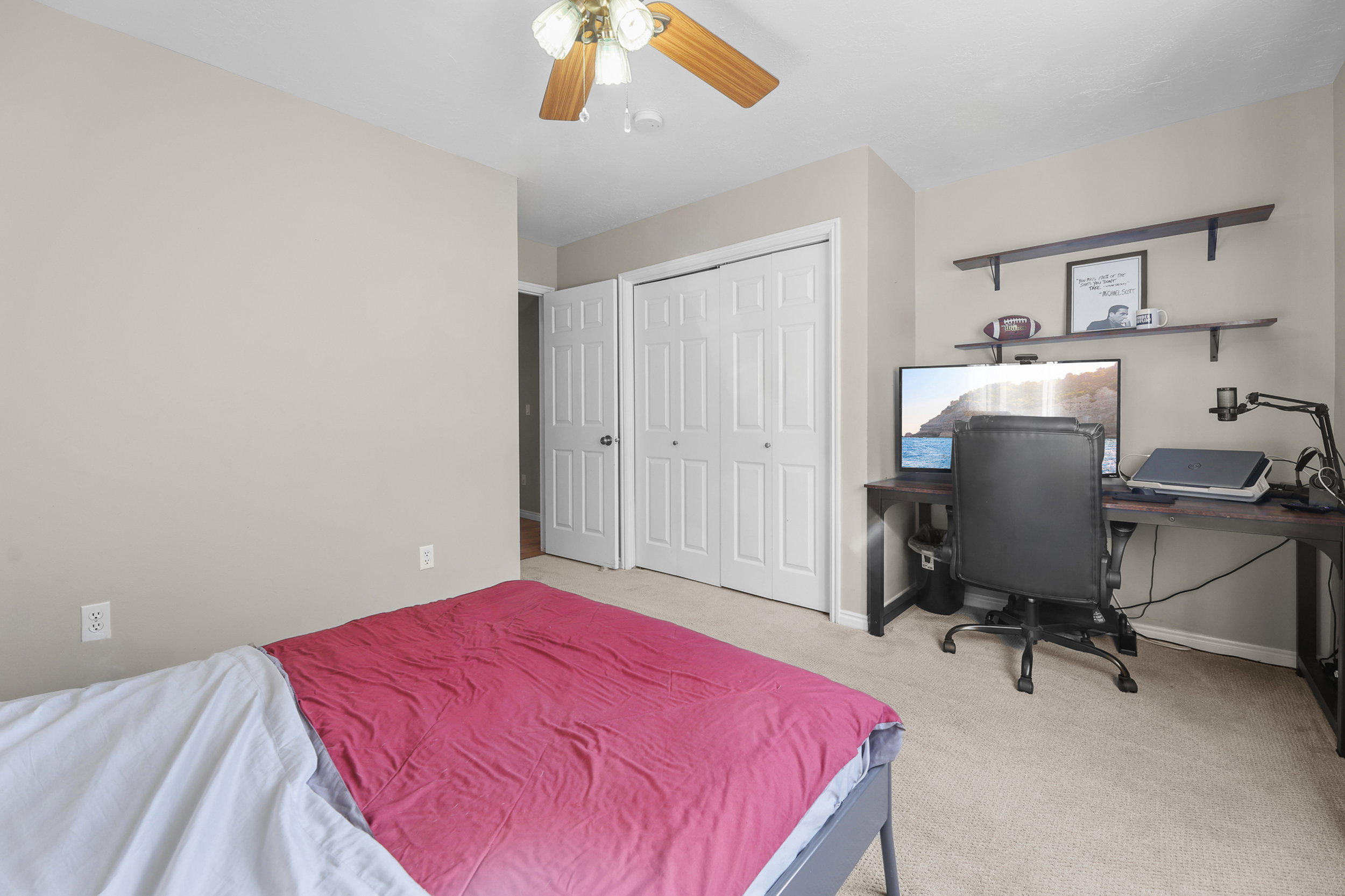 Carpeted room with a ceiling fan featuring wood-finish blades, a double-door closet with louvered panels, and a built-in wall shelf system