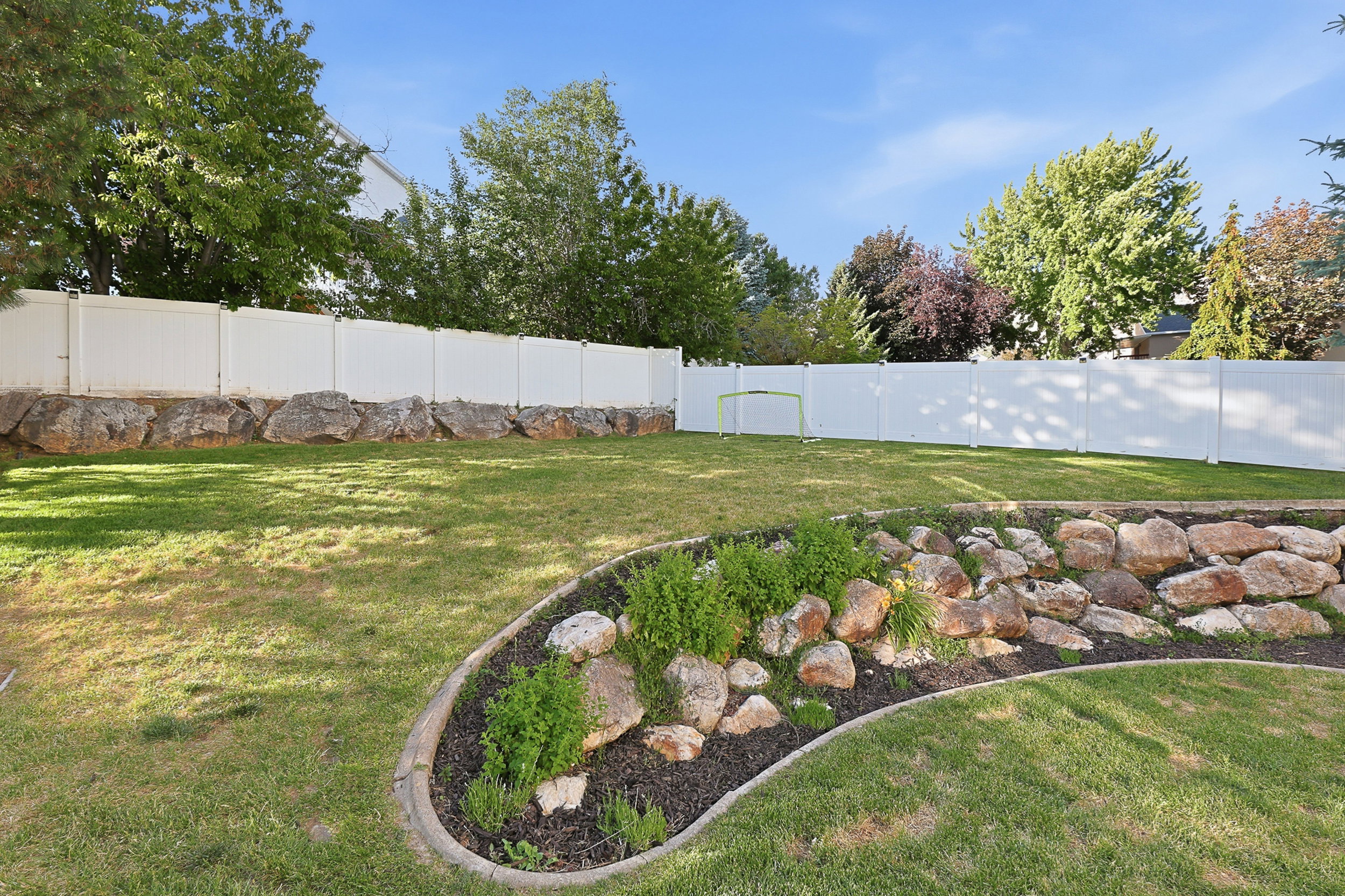 Expansive backyard featuring a manicured lawn, a white privacy fence, and a rock-lined garden bed with plantings