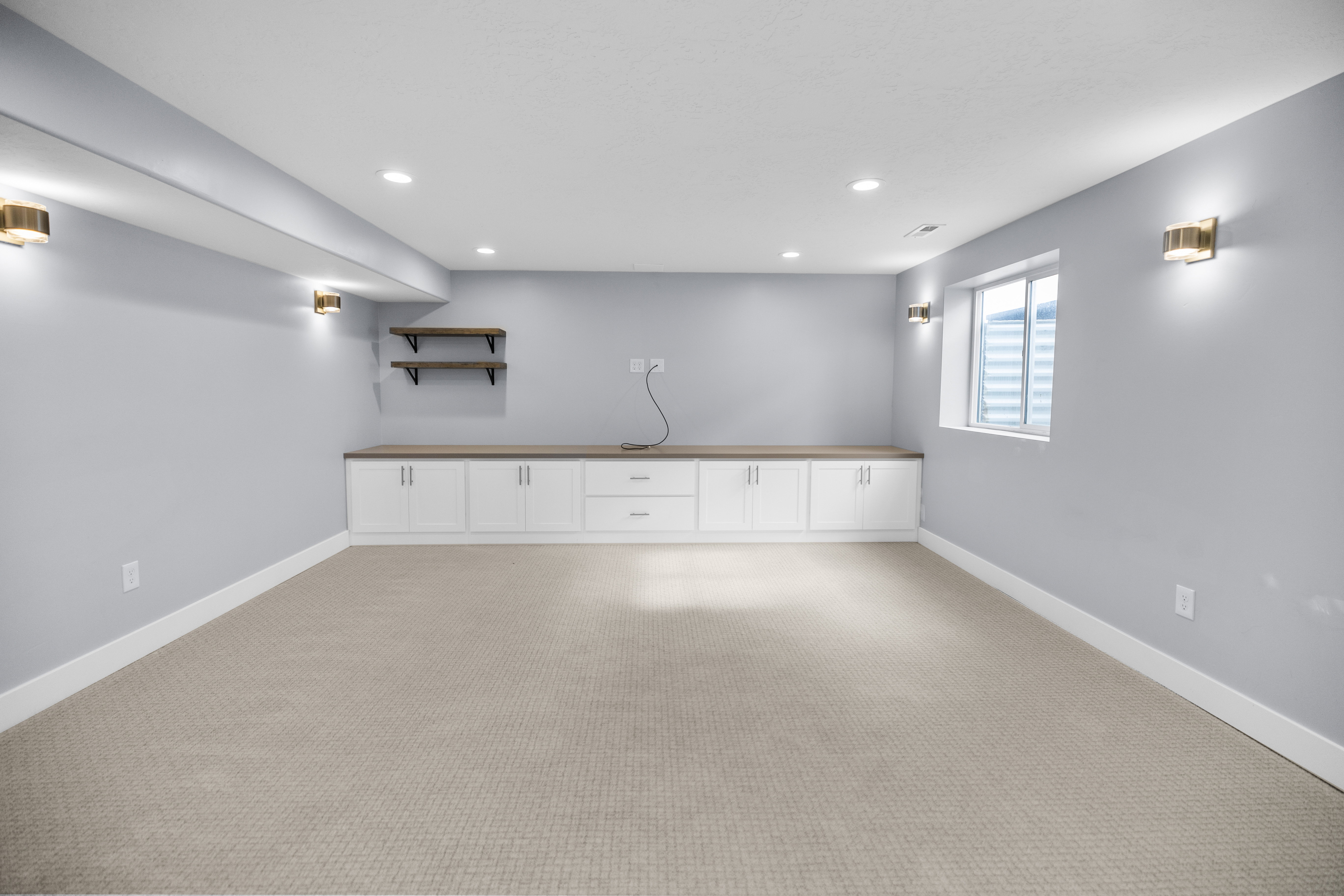 Spacious carpeted room featuring a built-in white cabinetry unit with a wood-finish countertop, recessed lighting, wall sconces, and a window with a window well