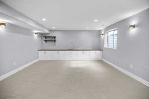 Spacious carpeted room featuring a built-in white cabinetry unit with a wood-finish countertop, recessed lighting, wall sconces, and a window with a window well