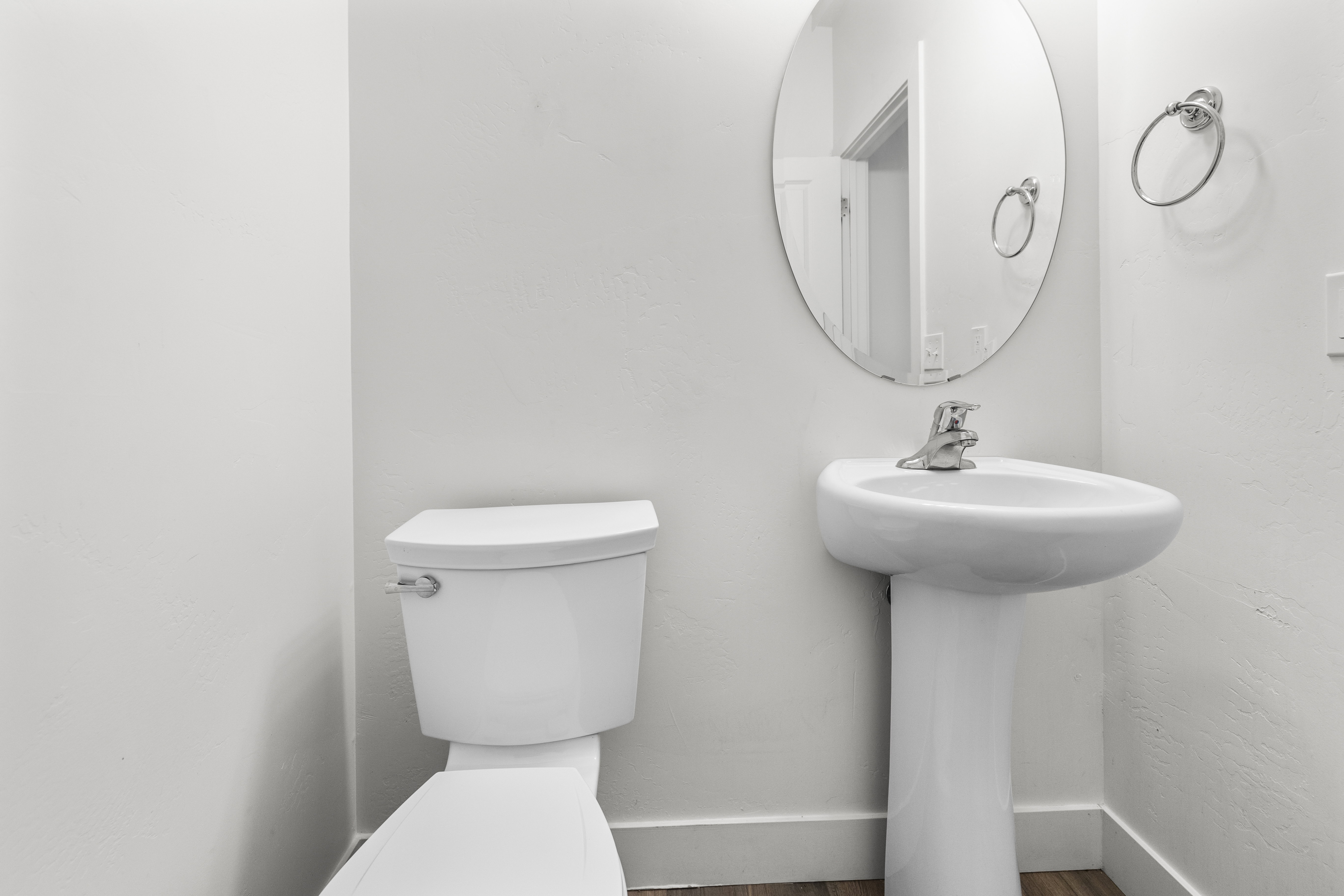 White pedestal sink with a chrome faucet, oval mirror, white toilet, and wood-finish flooring