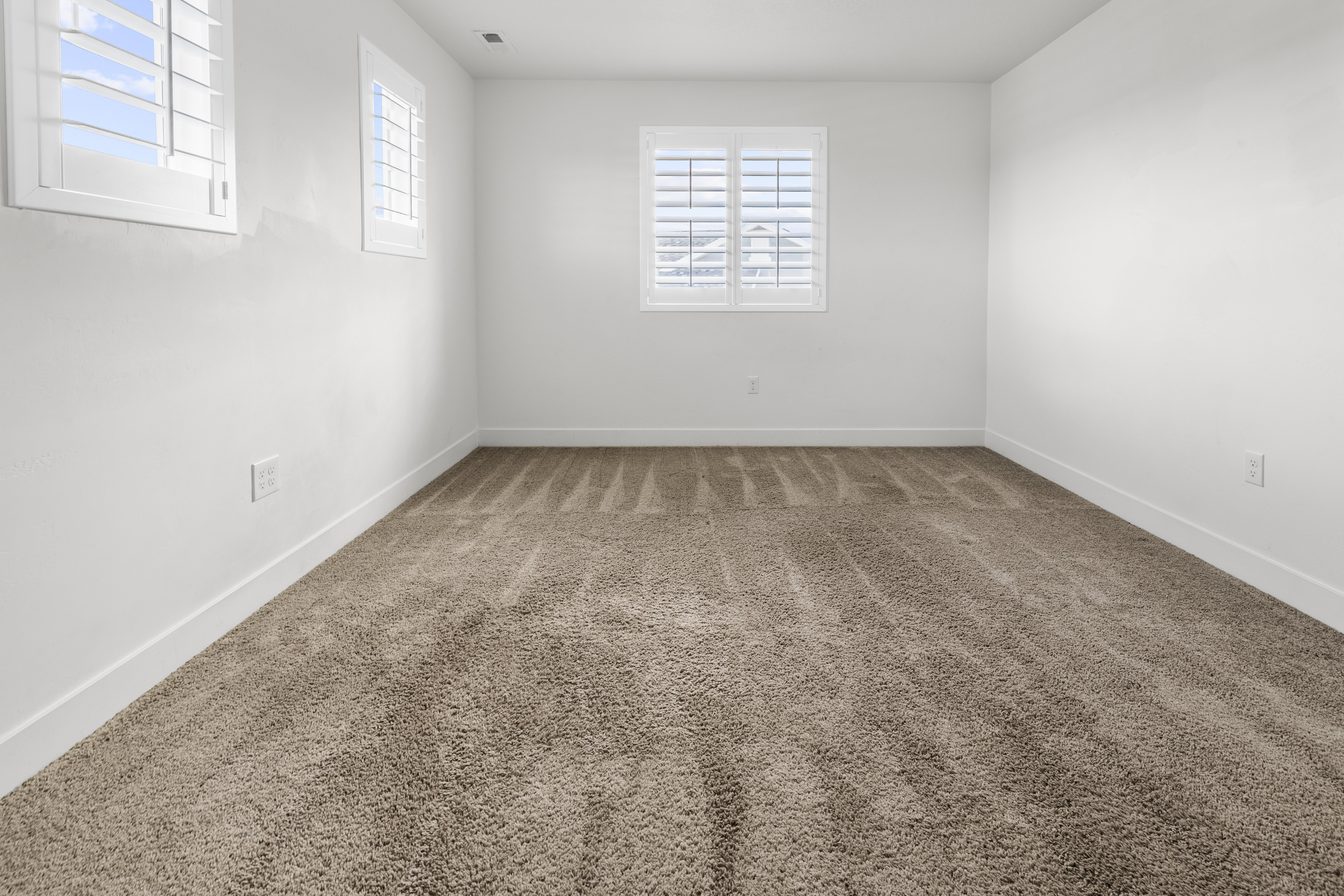 Bedroom 2. Bright room featuring two windows with white plantation shutters, white walls, and plush brown carpeting