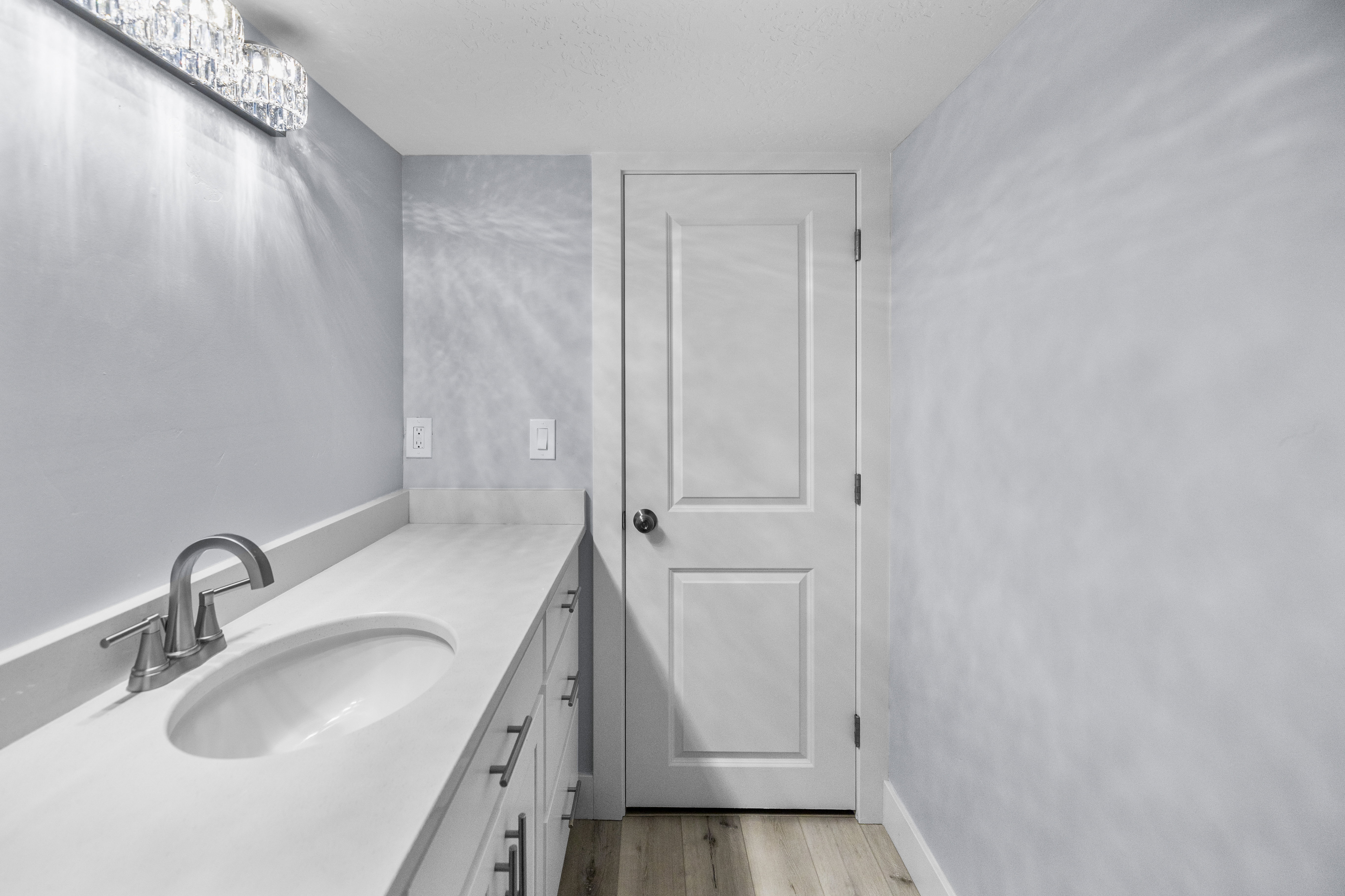 Bathroom vanity featuring an integrated sink, brushed nickel faucet, light-toned countertop, white cabinetry with dark hardware, and wood-finish flooring