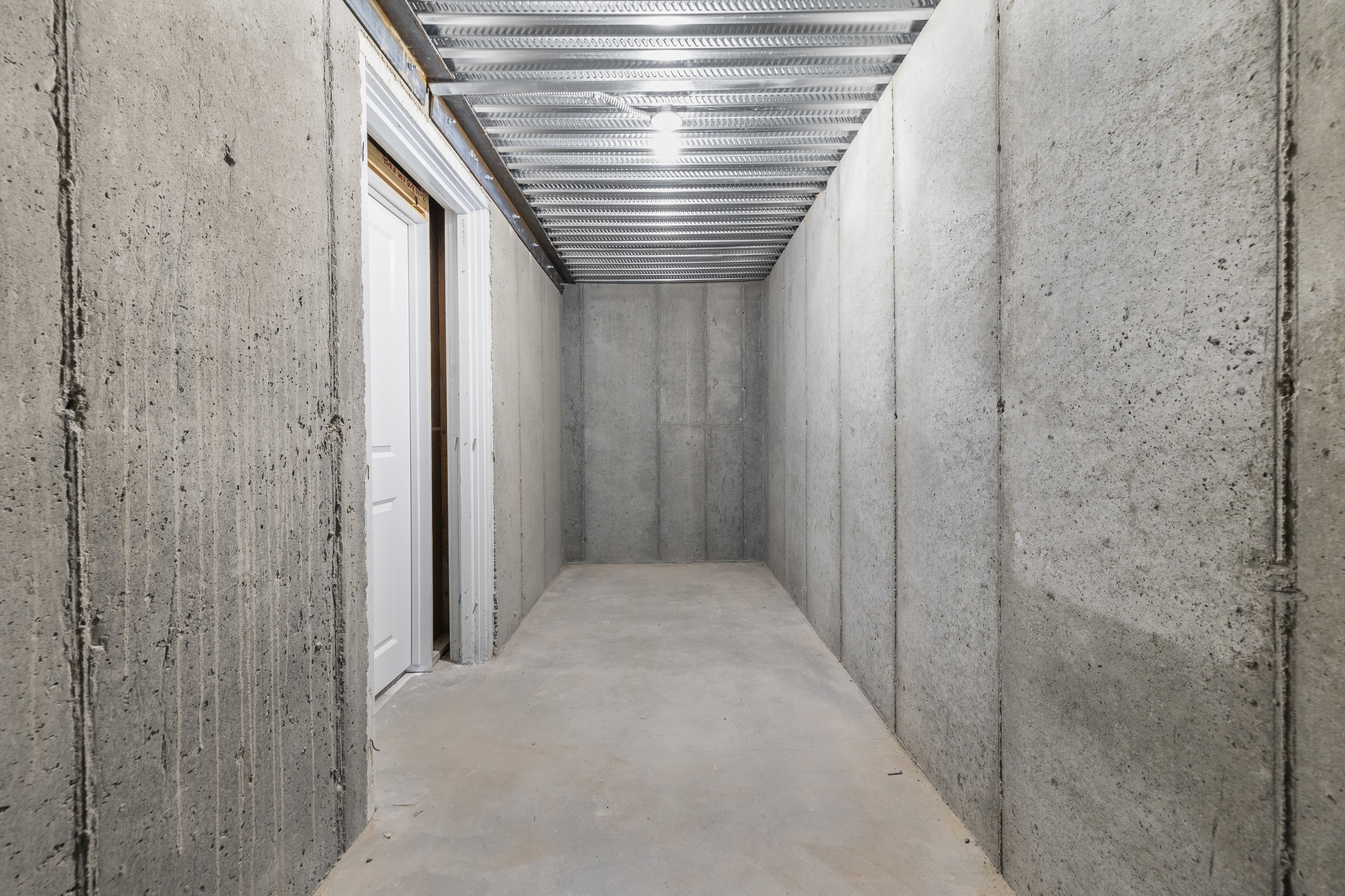 Cold storage space under the front porch in the basement featuring concrete slab flooring, poured concrete walls, exposed metal ceiling joists, and a white interior door frame