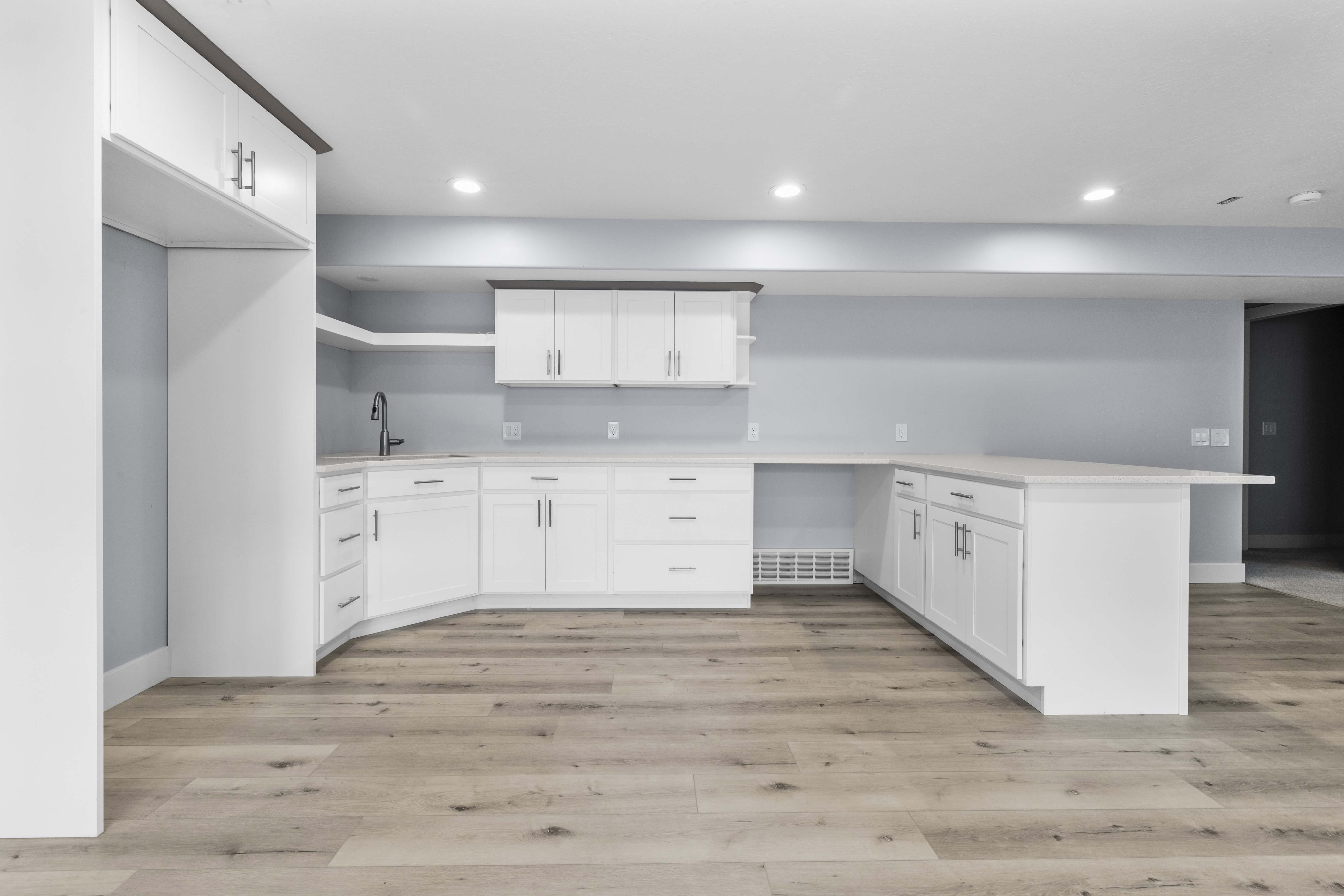 Contemporary kitchen area featuring white cabinetry, light-toned wood-finish flooring, recessed lighting, a built-in sink with a matte black faucet, and light-colored countertops