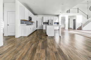 Open-concept living area featuring wood-finish flooring, a white kitchen island, white cabinetry, a dark subway tile backsplash, and a staircase with white balusters