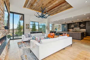 Living area featuring light wood-style flooring, a stone fireplace, expansive windows, suspended lighting, and a wood tray ceiling