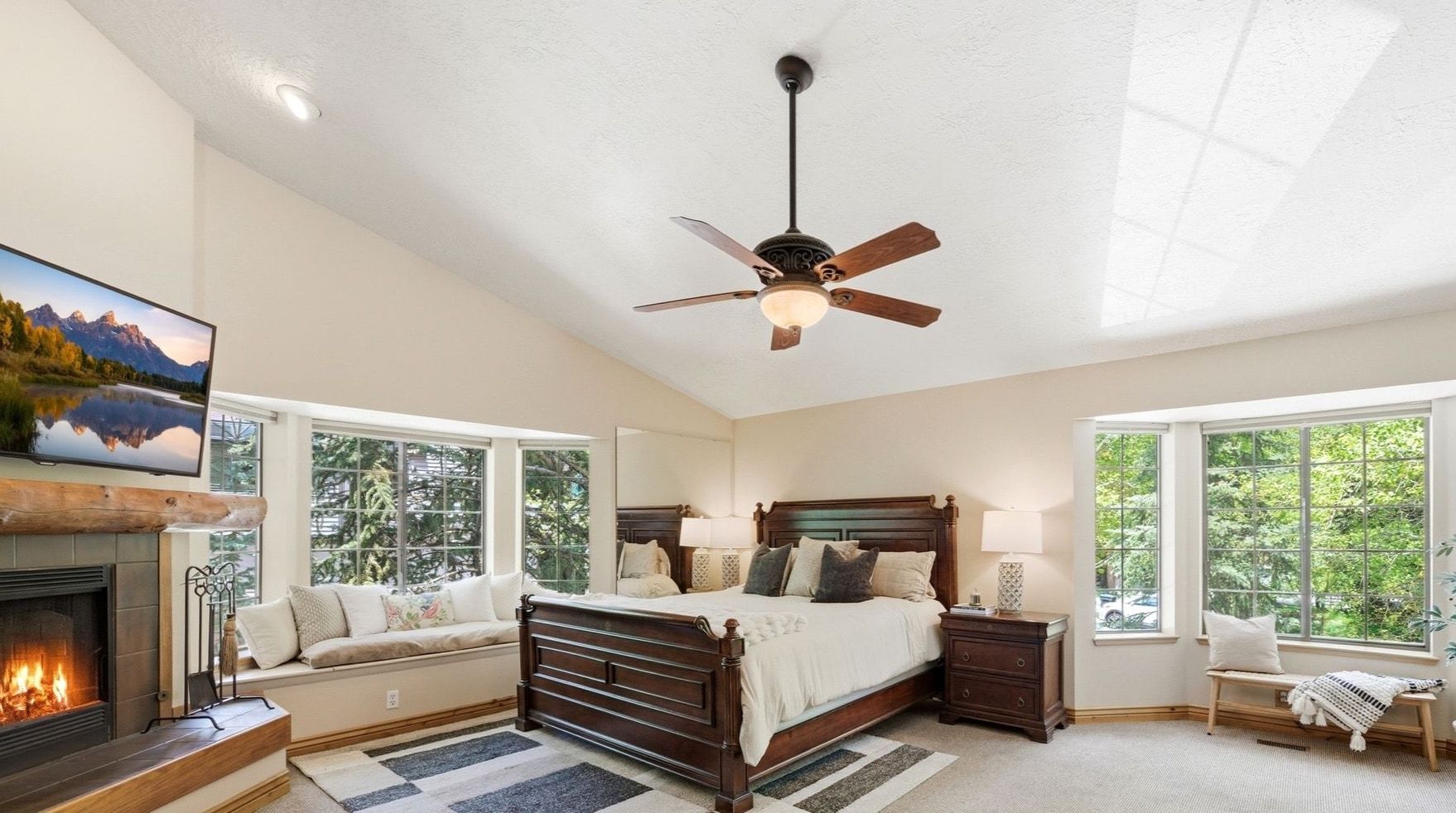 Vaulted ceiling room featuring a wood-burning fireplace with a log mantel, a built-in window seat, a ceiling fan, and large windows