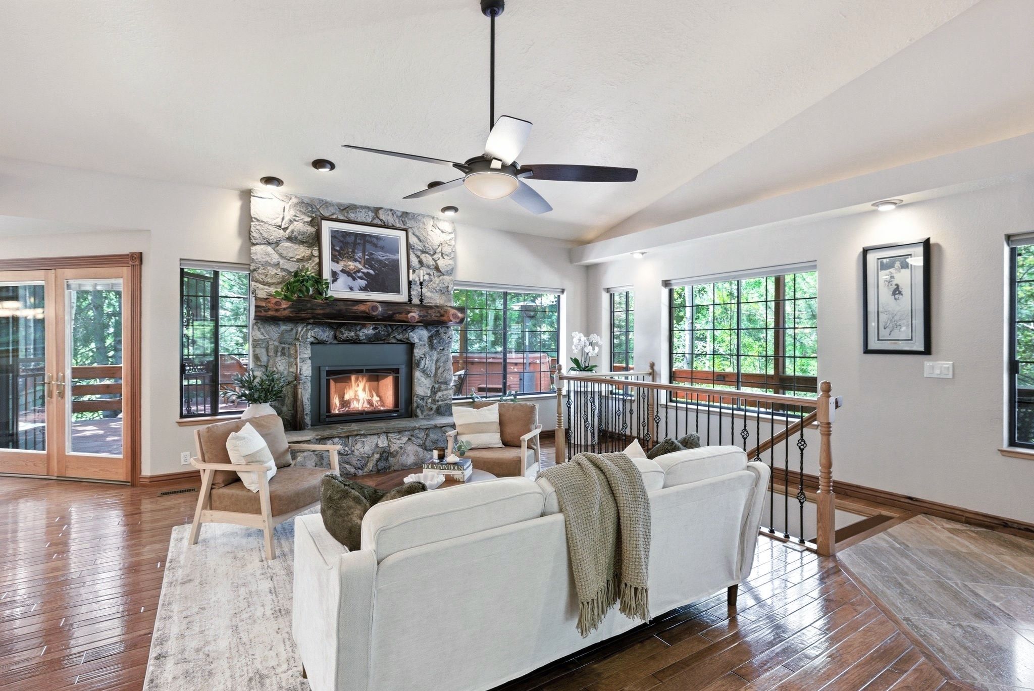 Spacious living area featuring a stone-faced fireplace with a substantial timber mantel, wood-finish flooring, a vaulted ceiling, and multiple windows