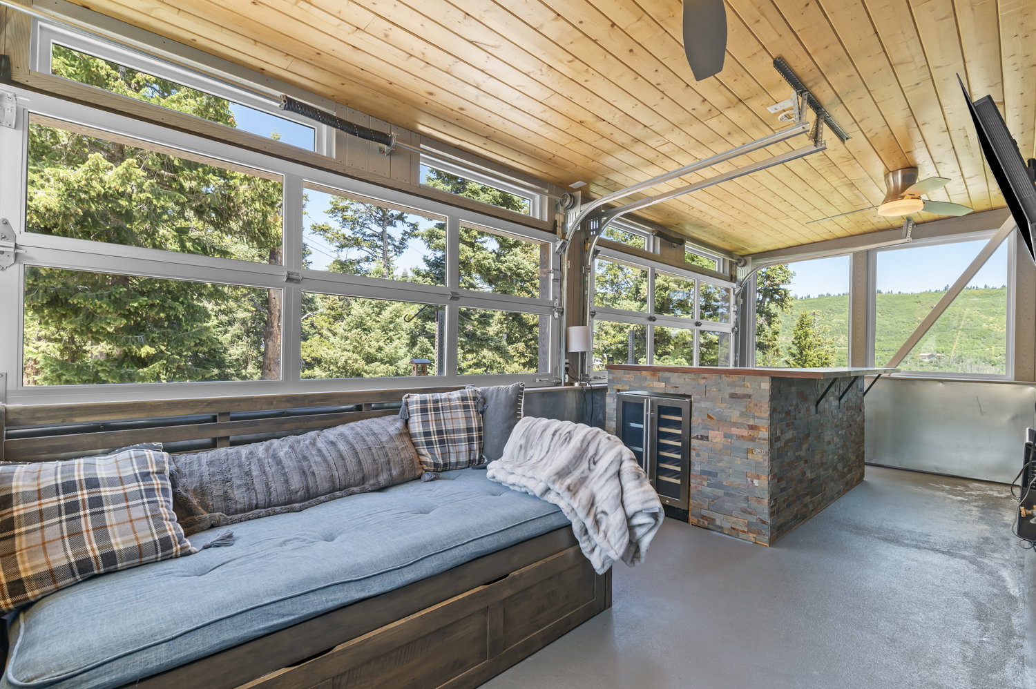 Indoor-outdoor living space featuring a full-width glass garage door, wood-paneled ceiling, stone-clad bar with built-in beverage coolers, and durable flooring