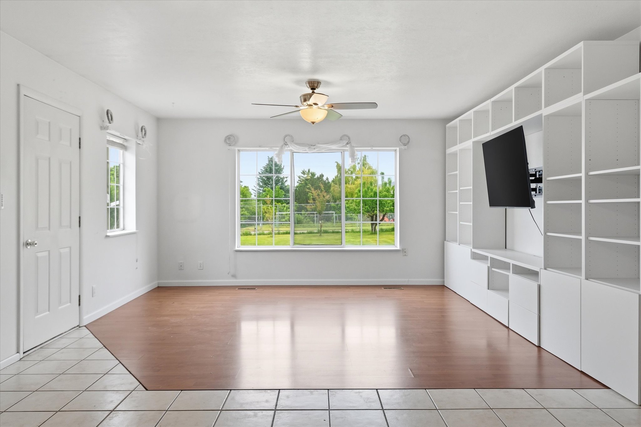 Spacious interior featuring a large picture window, wood-finish flooring, a built-in shelving system with integrated media display, and a ceiling fan