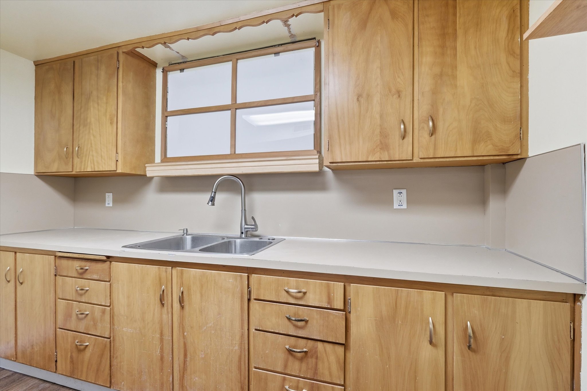 Kitchen featuring wood-finish cabinetry, a double basin stainless steel sink with a gooseneck faucet, light-toned countertops, and a window with wood framing