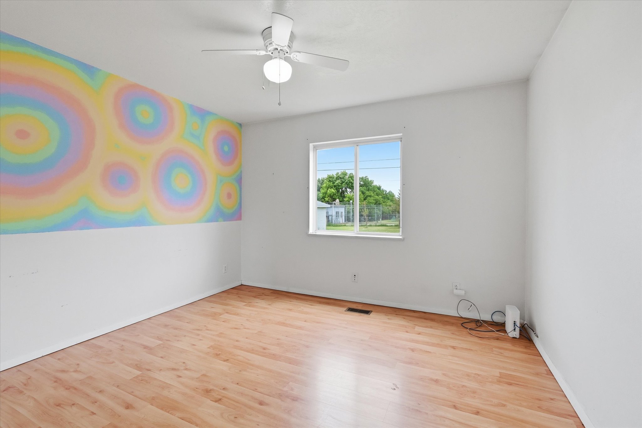 Spacious room featuring wood-finish flooring, a contemporary ceiling fan with integrated lighting, a large window providing outdoor visibility, and bright white walls with a unique accent wall design