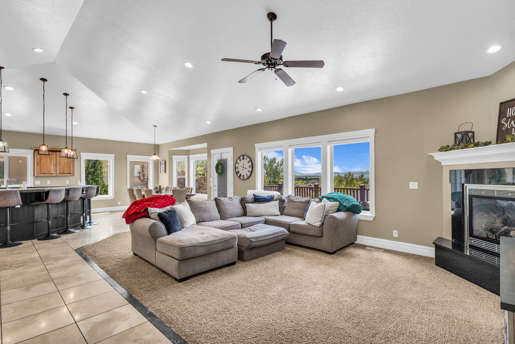 Open-concept living area featuring vaulted ceilings, recessed lighting, a ceiling fan, a built-in fireplace with a mantel, and multiple windows offering expansive outdoor scenery