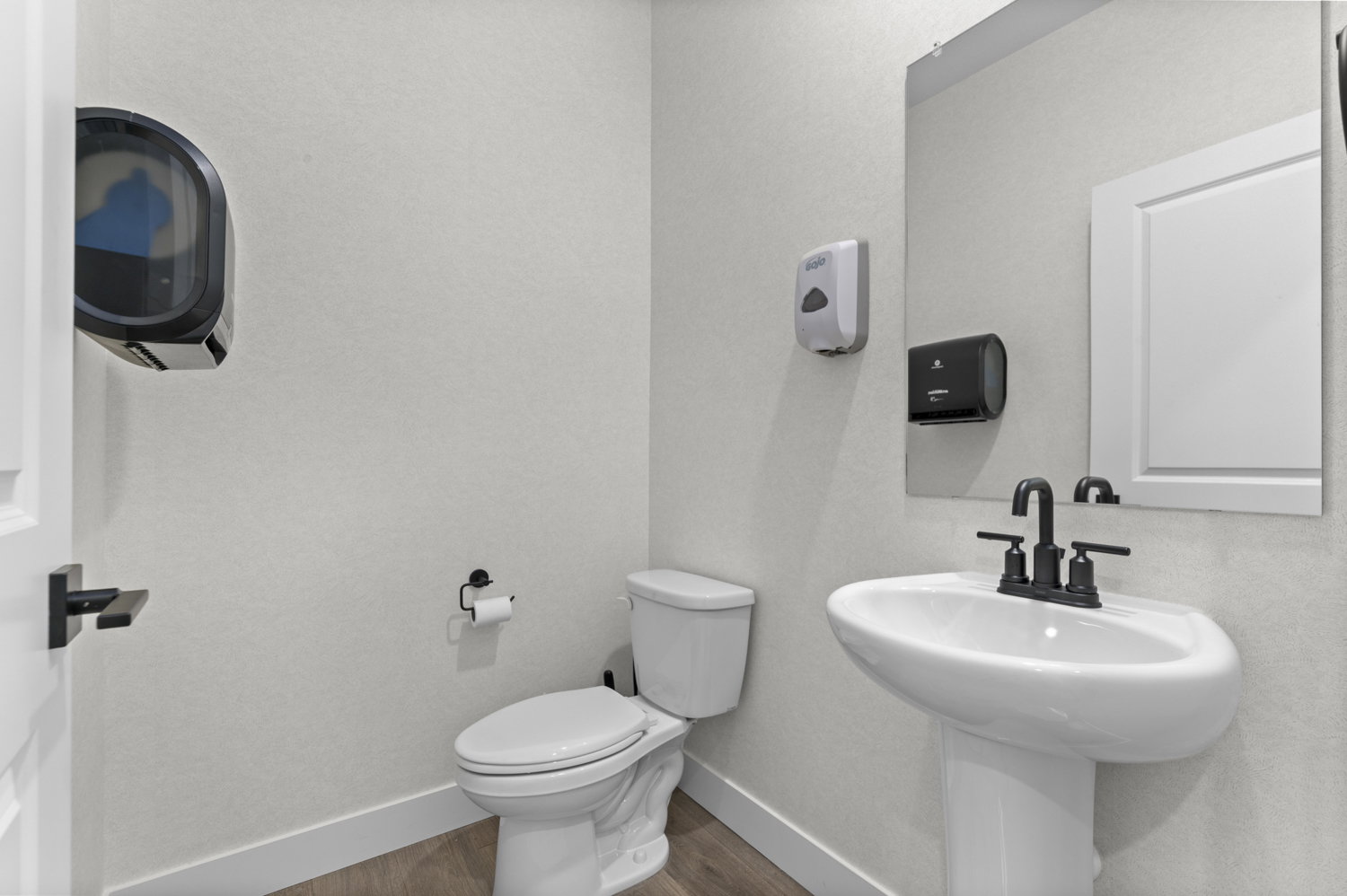 White pedestal sink with matte black faucet, wall-mounted mirror, white commode, and wood-finish flooring