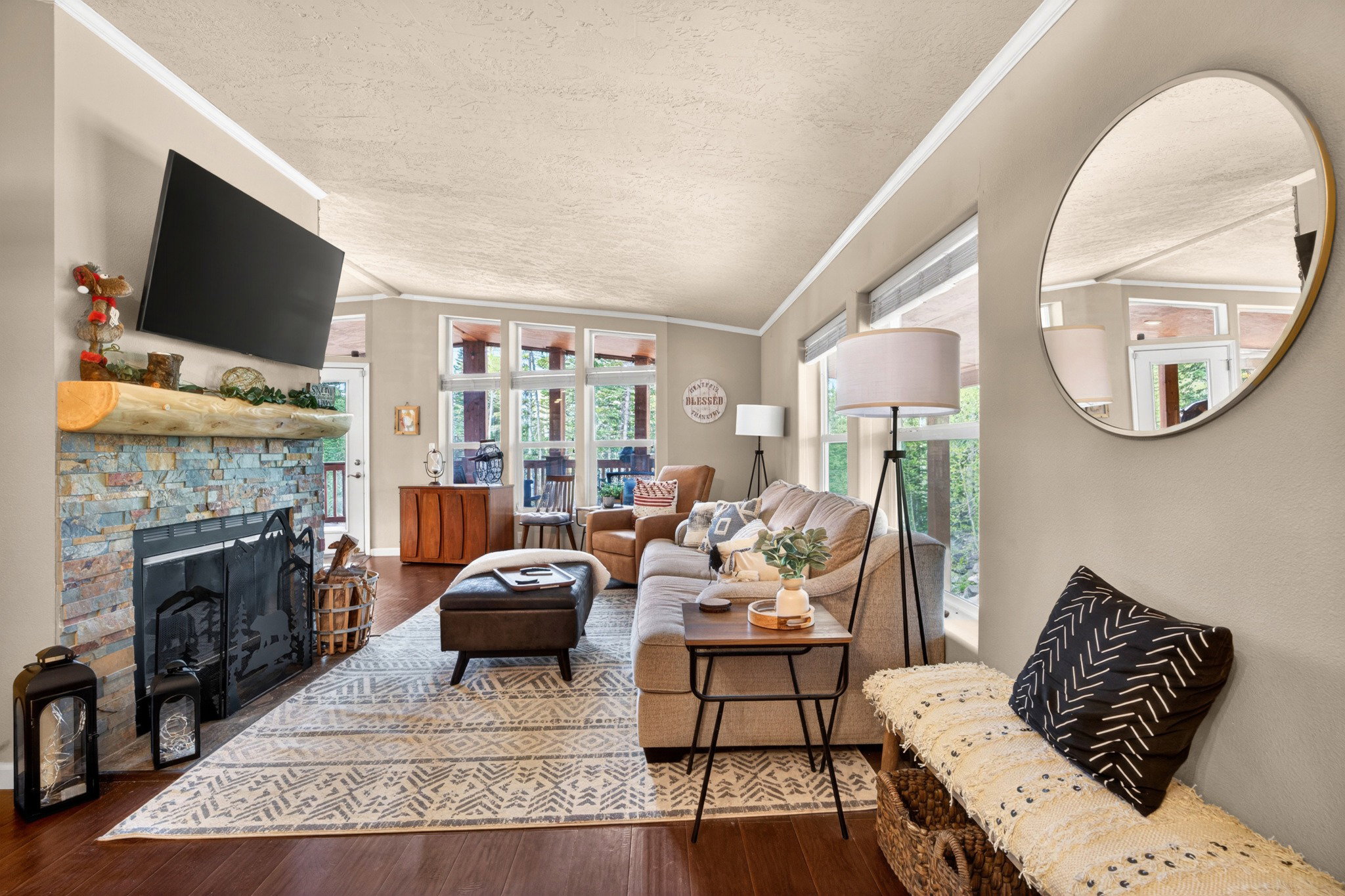 Living area featuring wood-finish flooring, a stacked stone fireplace with a substantial timber mantel, and a wall of windows