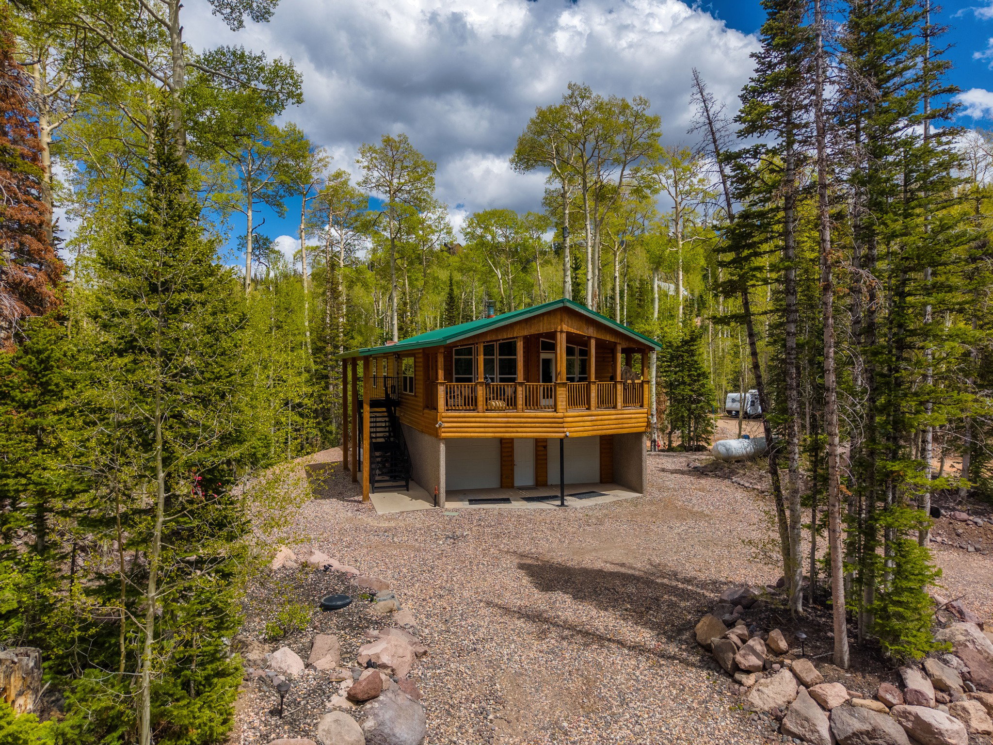 Log cabin exterior with a green metal roof, elevated deck, full-length windows, and surrounding mature trees