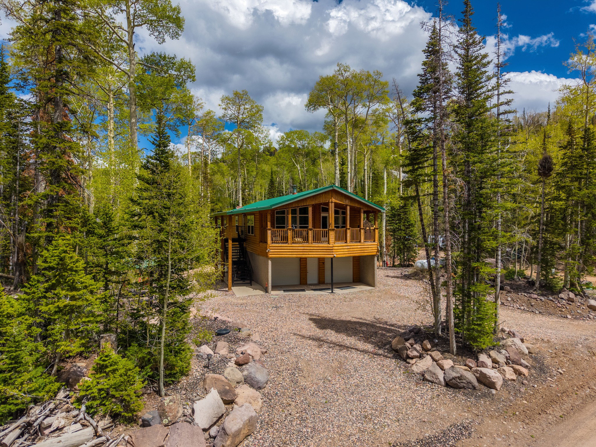 Log-sided cabin with a green metal roof, elevated on concrete stilts, featuring a wraparound wood-plank deck with a railing, and a gravel driveway surrounded by mature trees