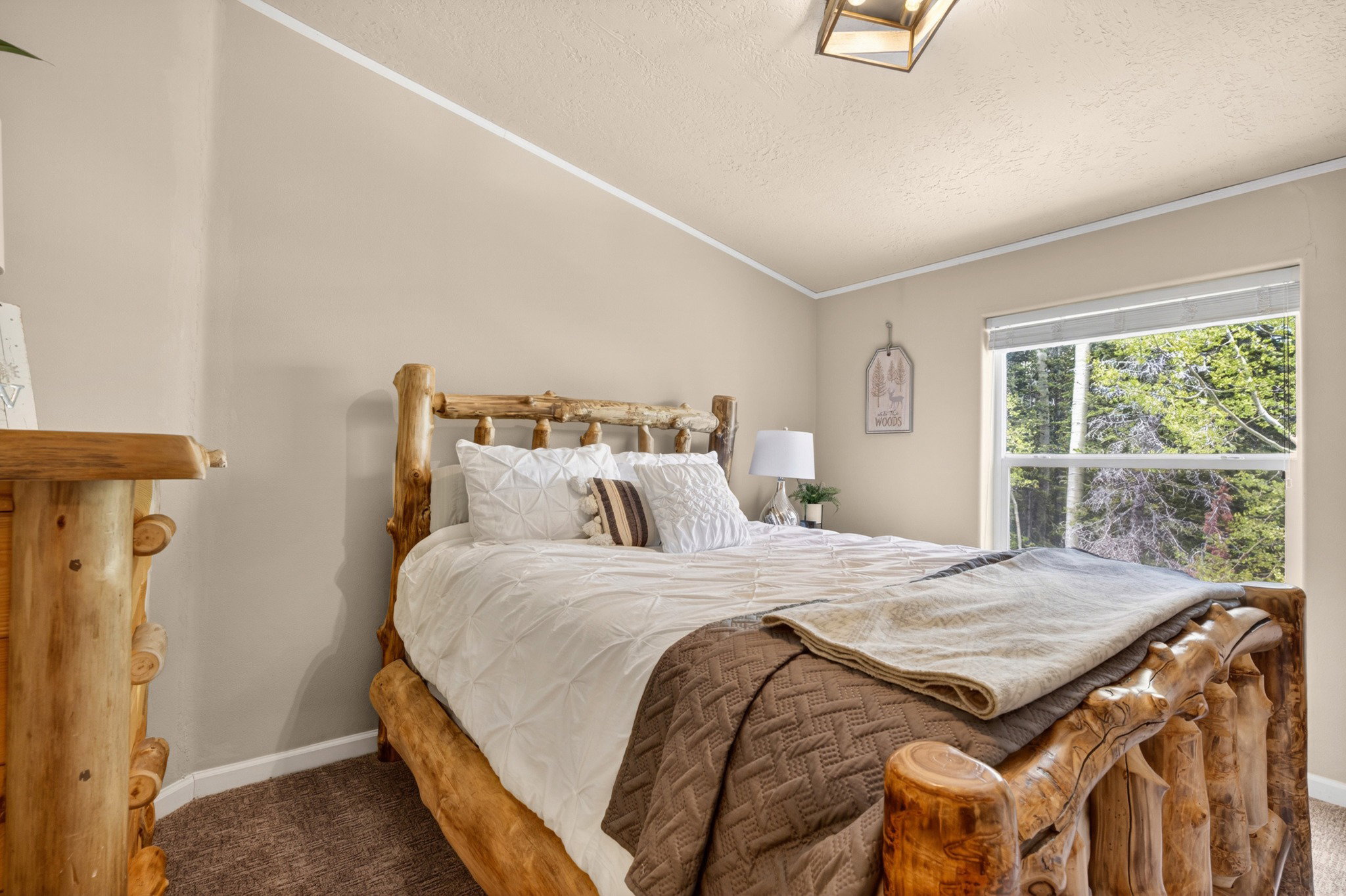 Spacious bedroom featuring natural wood bed frame and dresser, window with verdant tree scenery, neutral-toned walls, and recessed lighting