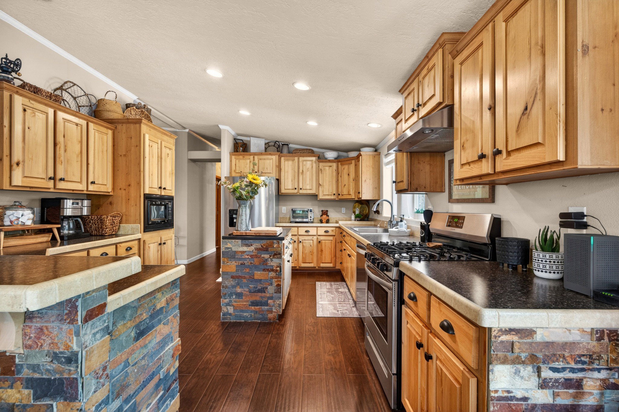 Spacious kitchen featuring extensive natural wood cabinetry, dark stone-finish countertops, stainless steel appliances, and wood-finish flooring