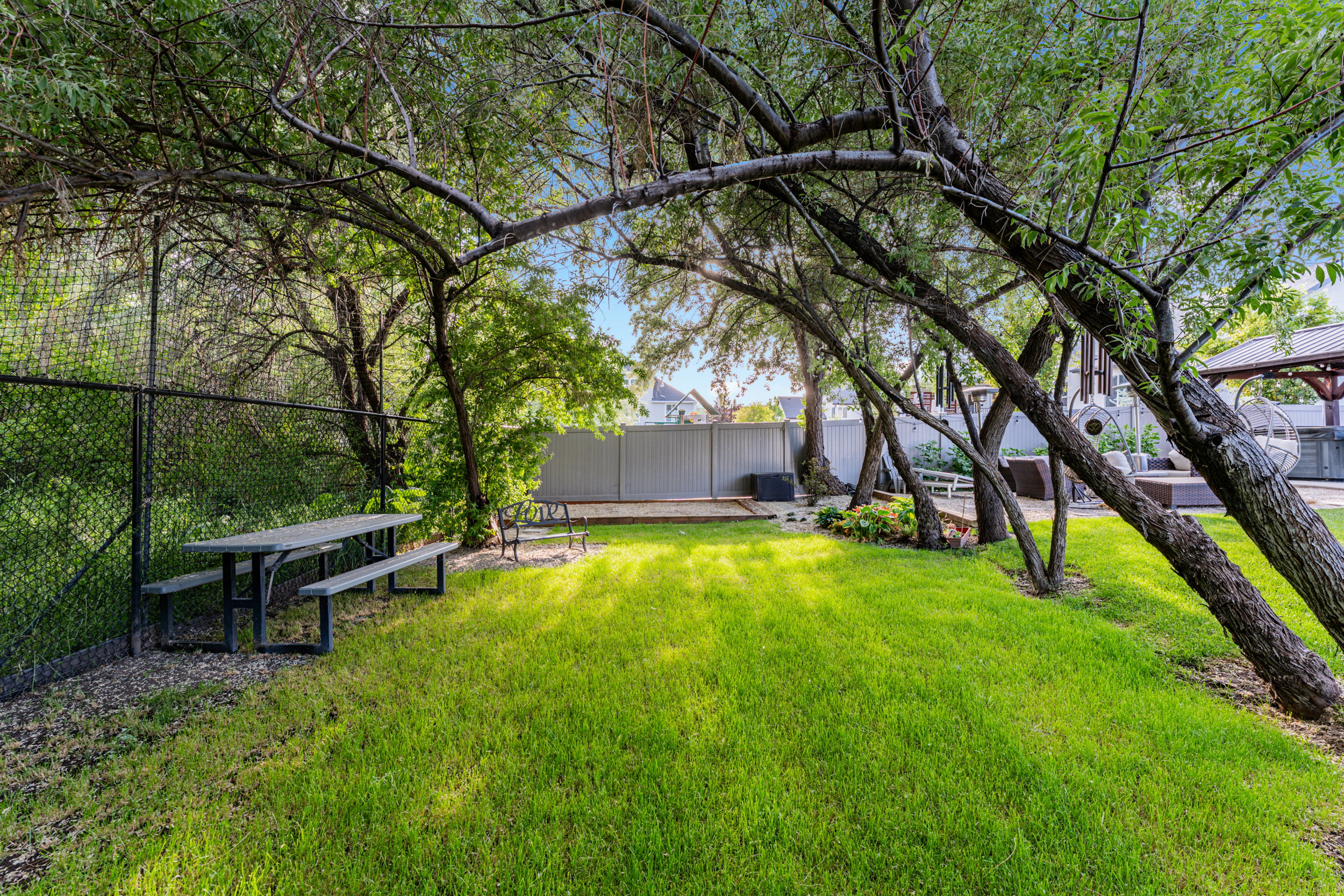 Expansive green space featuring mature trees, a picnic table, and a chain-link fence