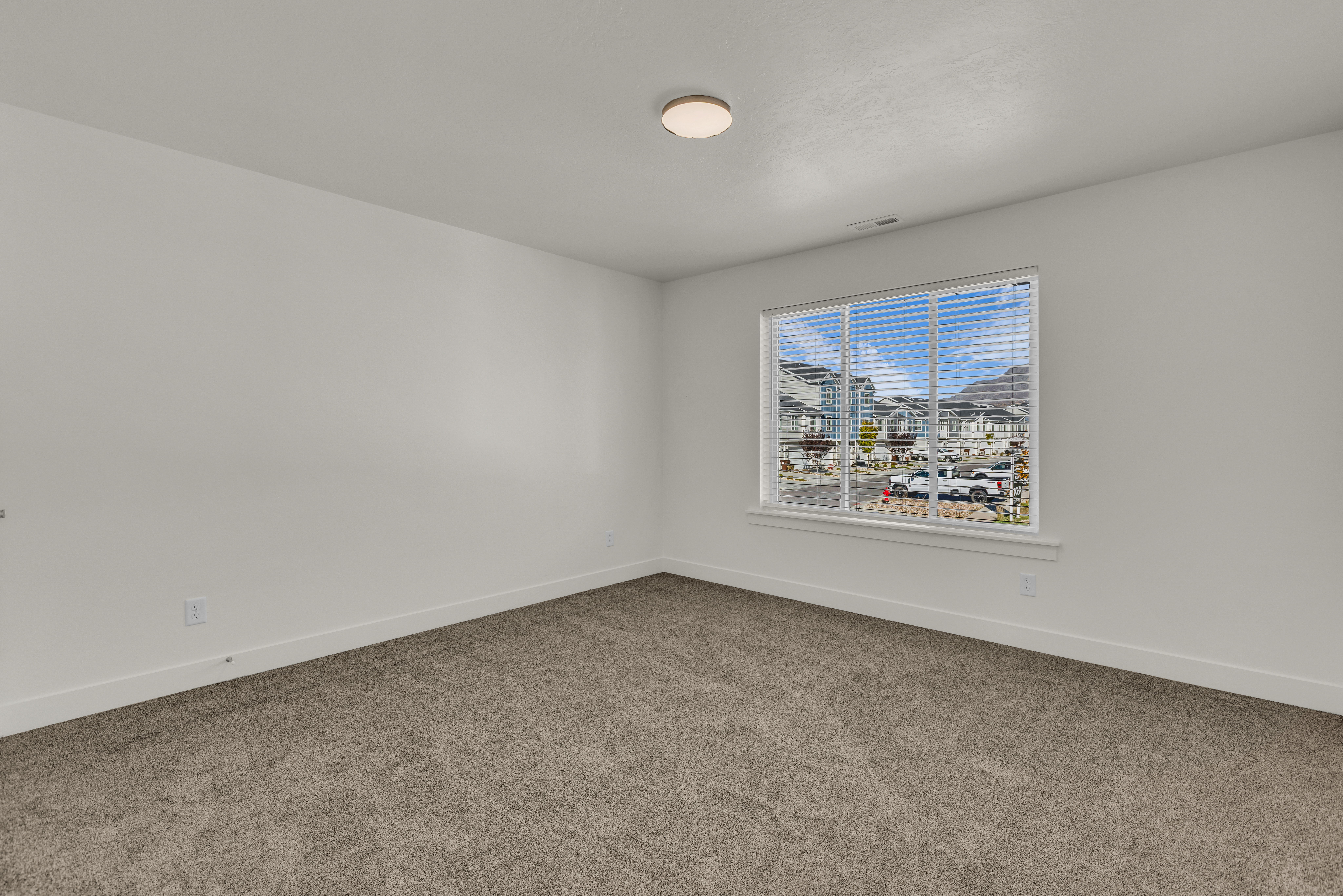 Spacious room featuring neutral carpeting, white painted walls, a large window with blinds, and an overhead flush-mount light fixture