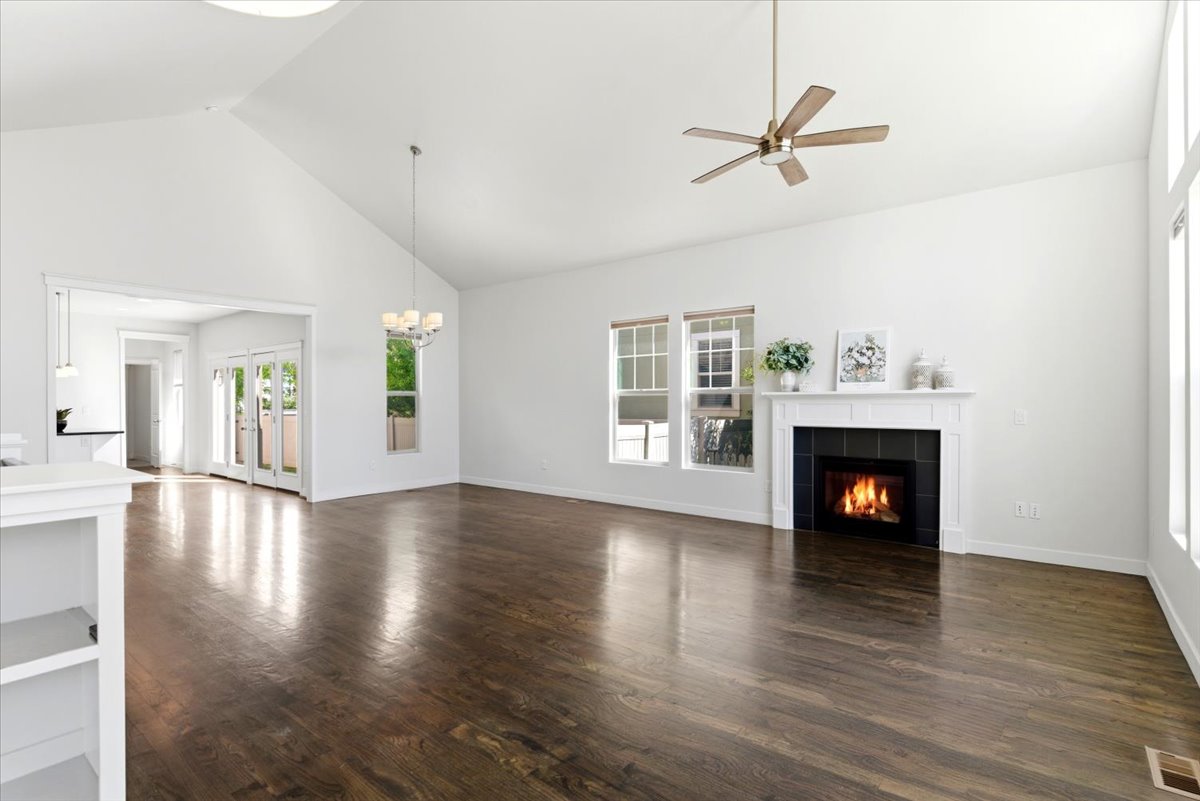 Spacious living room featuring vaulted ceilings, hardwood flooring, a fireplace with a white mantel and black tile surround, multiple windows, and a ceiling fan