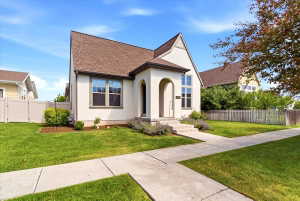 Stucco exterior with a peaked roofline, arched entry, mature landscaping, concrete sidewalk, and a privacy fence