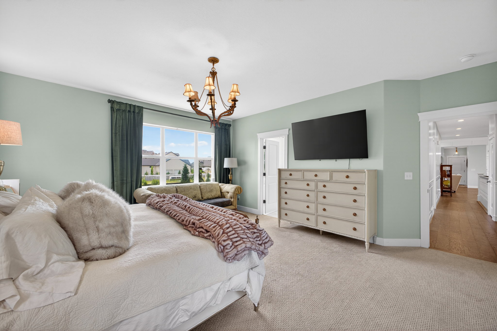 Spacious room with a large window, a classic chandelier, light green walls, light-colored carpeting, and a visible hallway with wood-finish flooring