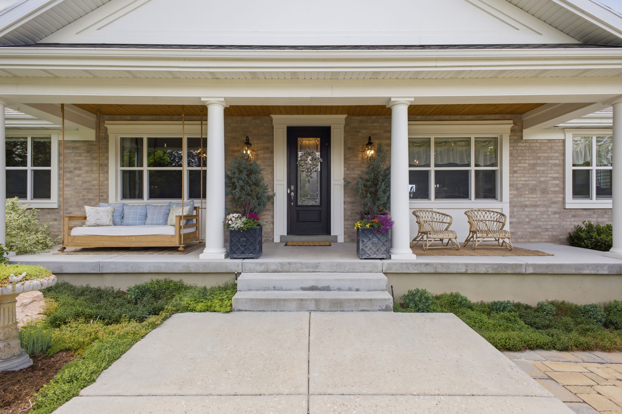 Inviting covered front porch with brick facade, substantial white columns, a dark entry door with decorative glass, wood-finish ceiling, and mature shrubbery flanking the concrete walkway