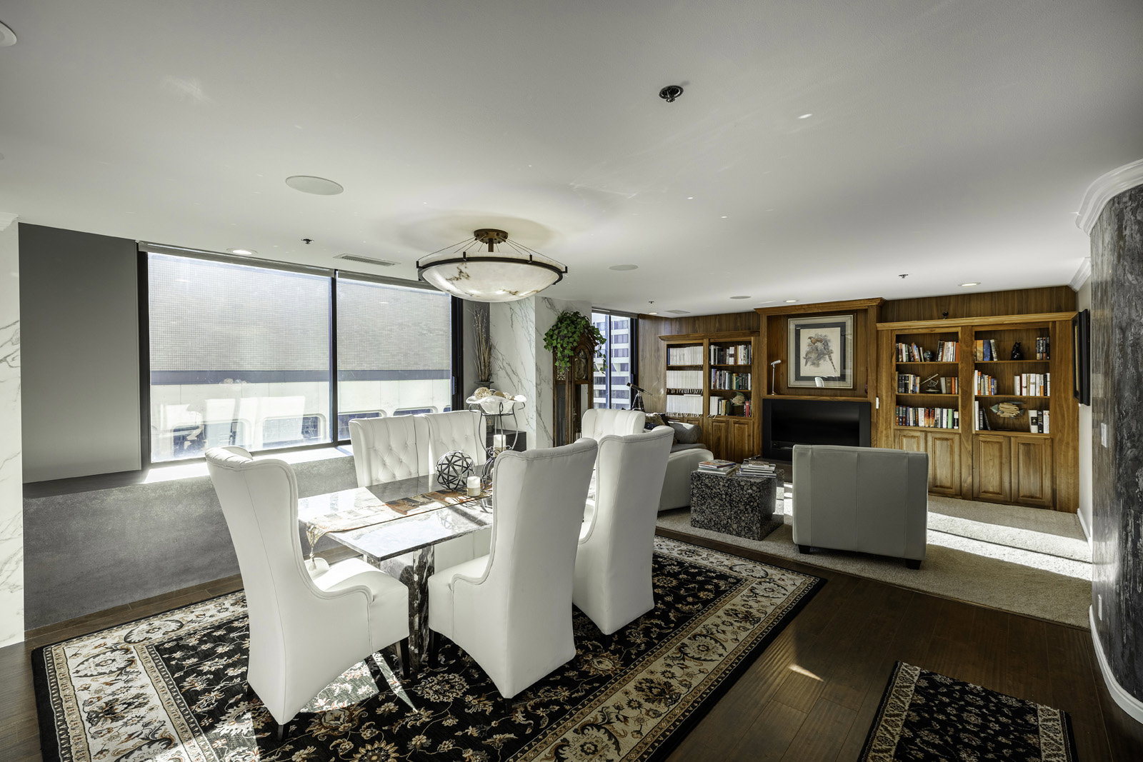 Open-concept living and dining area featuring wood-finish flooring, large windows with roller shades, a built-in fireplace with a dark surround, custom wood built-in shelving, and recessed lighting