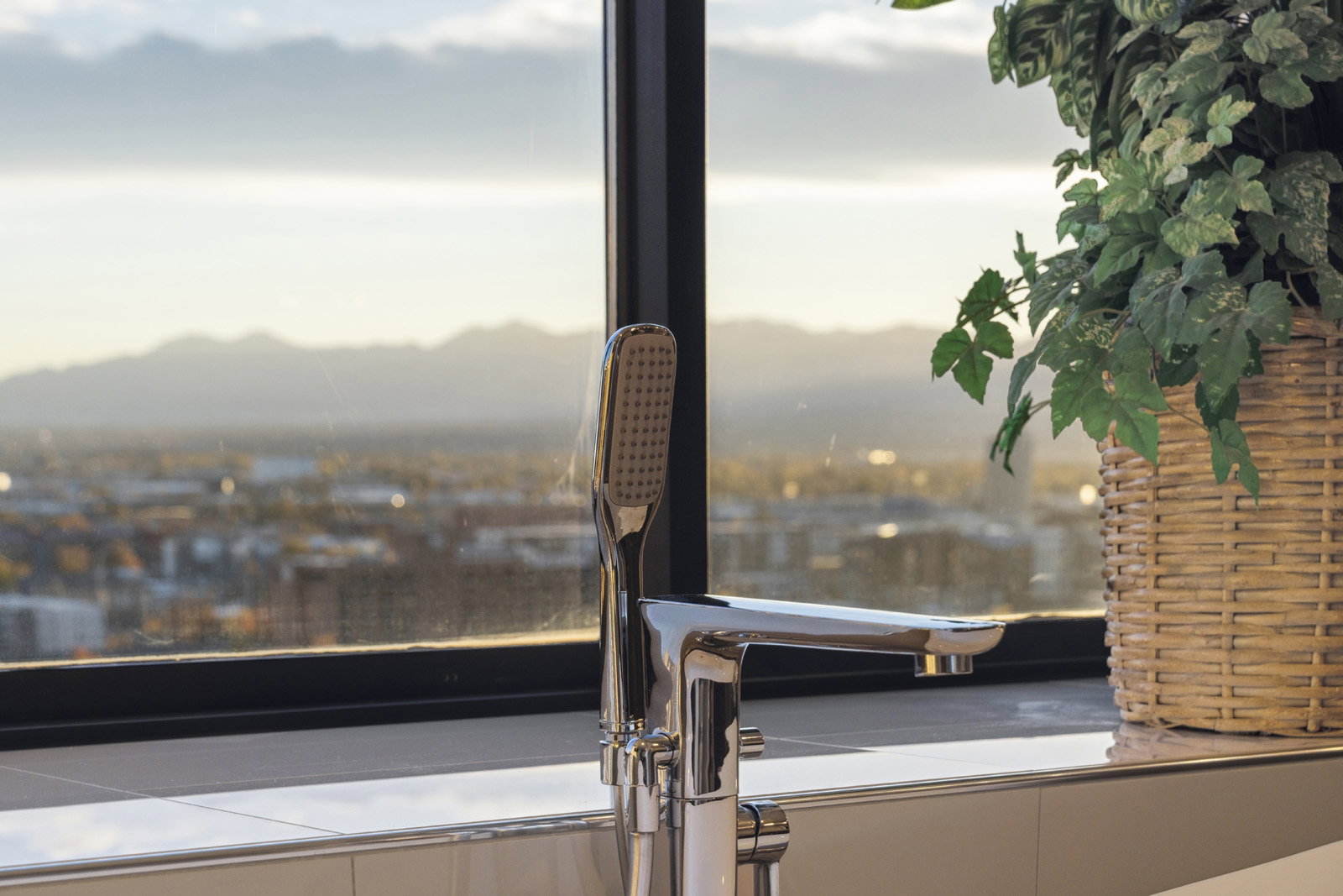 Modern chrome freestanding tub filler with integrated handheld shower, positioned against a large window featuring city and distant mountain scenery