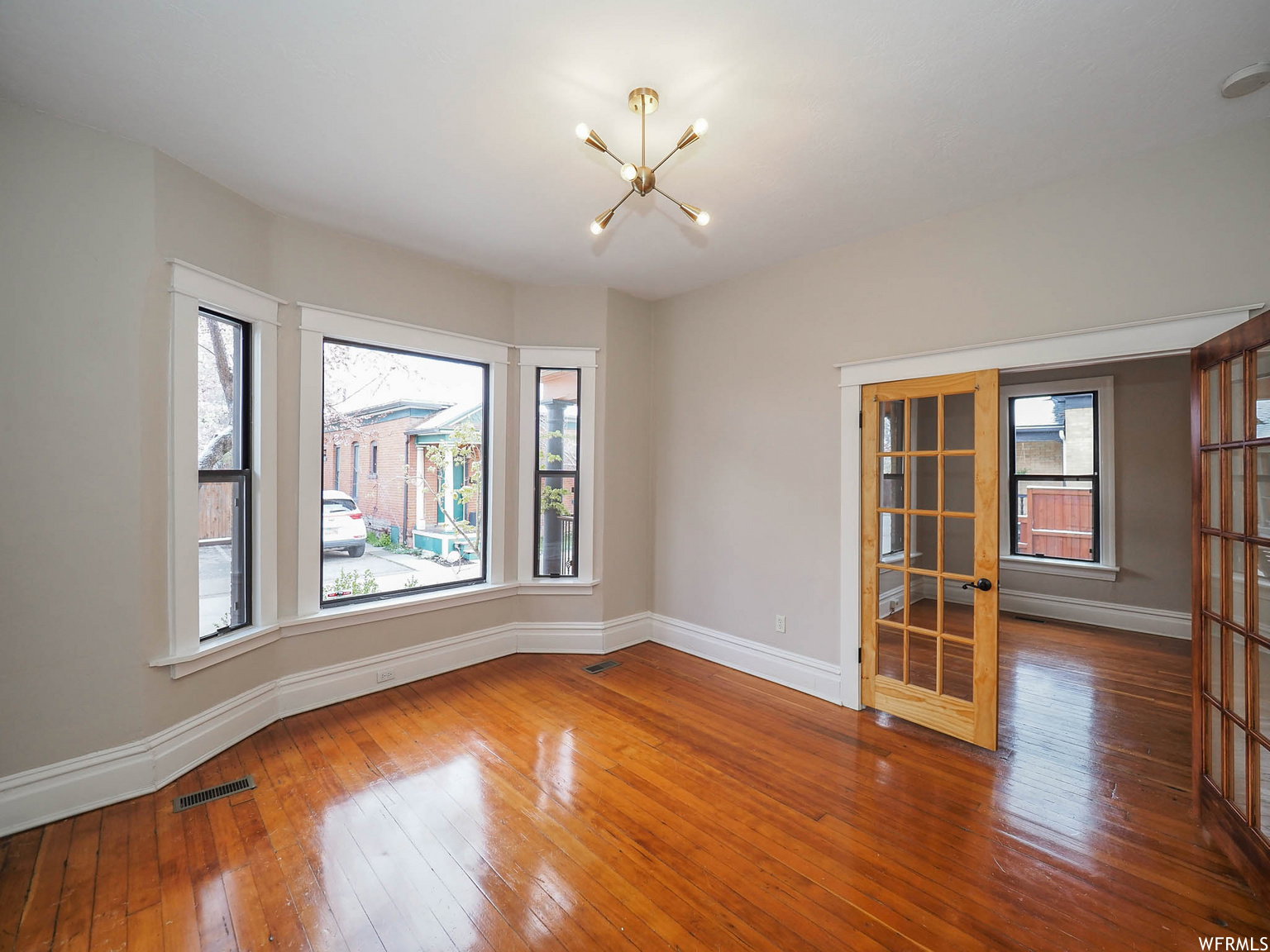 Hardwood flooring throughout featuring a bay window with white trim, a brass-finish sputnik light fixture, and two French doors with clear glass panes