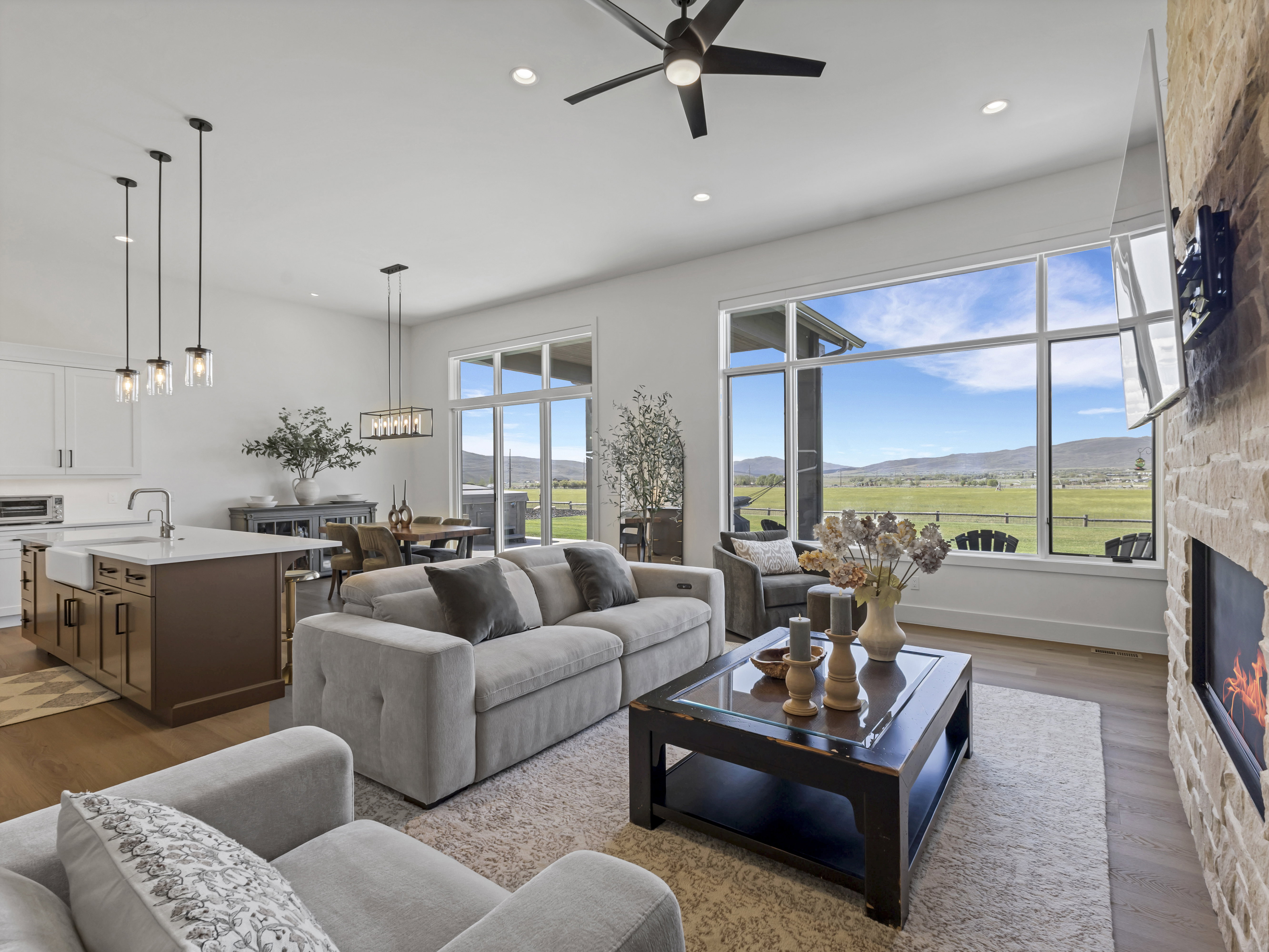Open-concept interior featuring light wood-finish flooring, a stone-clad fireplace, and expansive windows