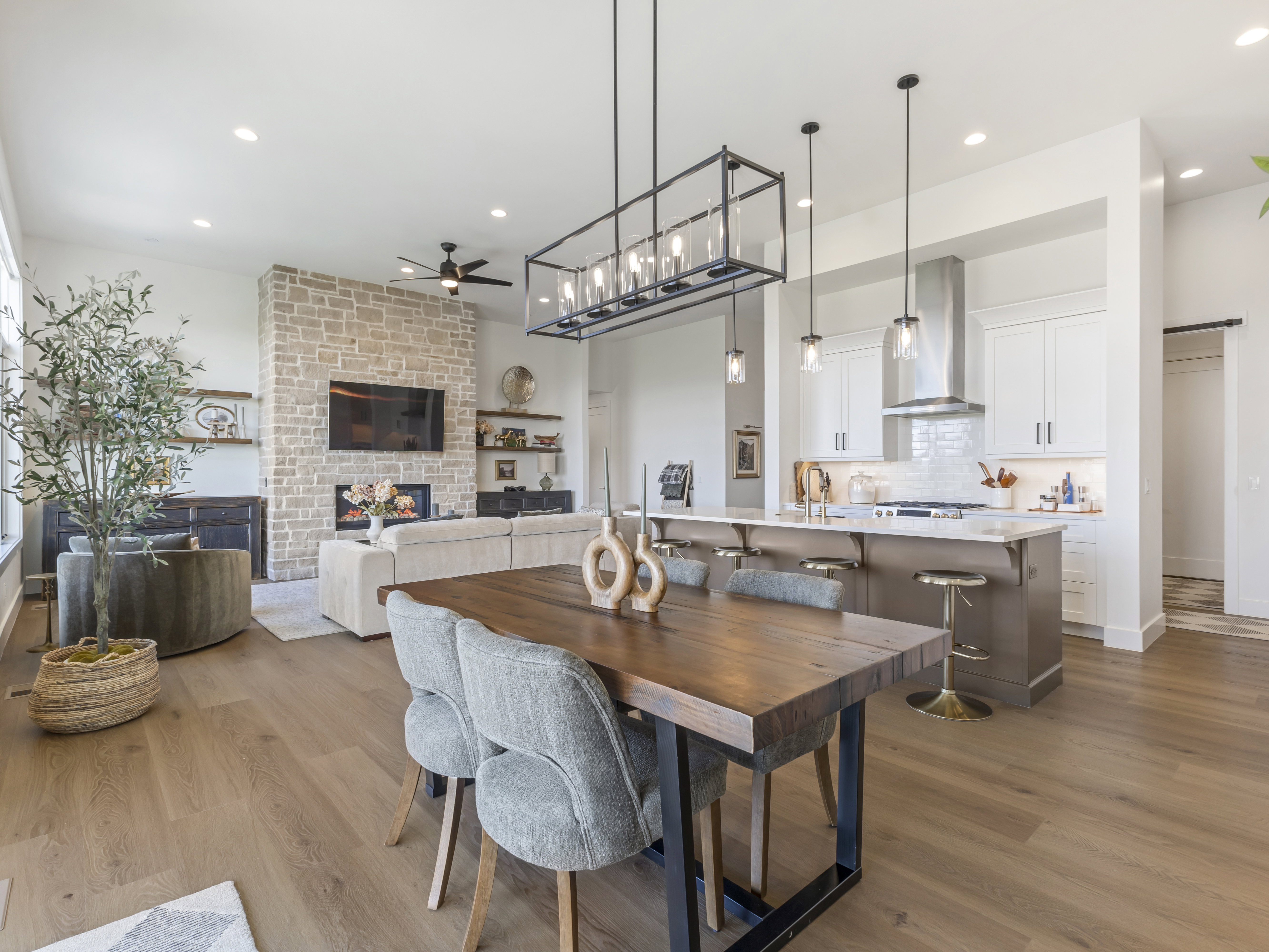 Open-concept living space featuring wood-finish flooring, a stone-clad fireplace with built-in shelving, a kitchen island with seating, pendant lighting, and recessed lighting