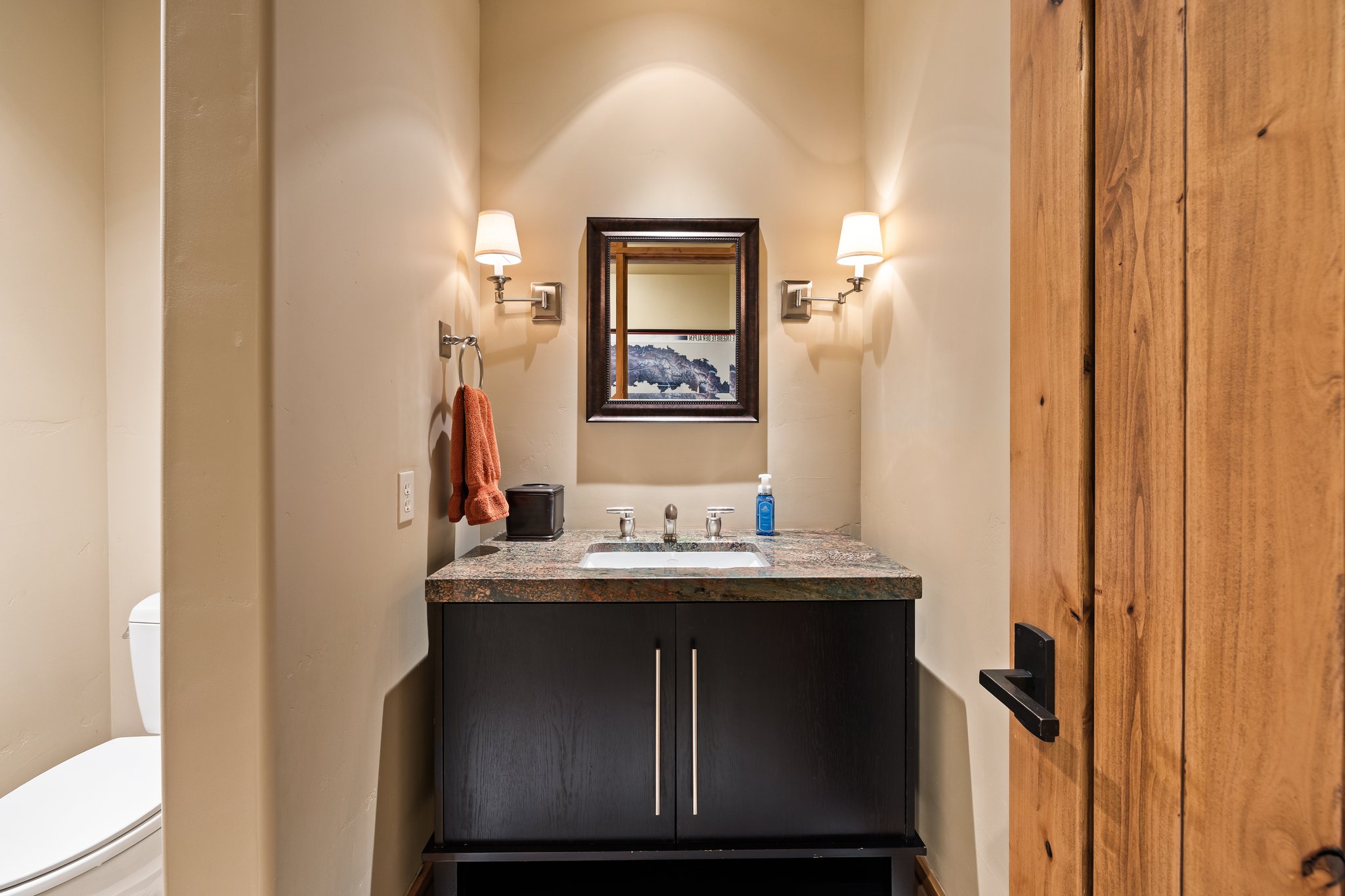 Powder room featuring a dark wood vanity with a stone-finish countertop and integrated sink, framed mirror, and wall sconces