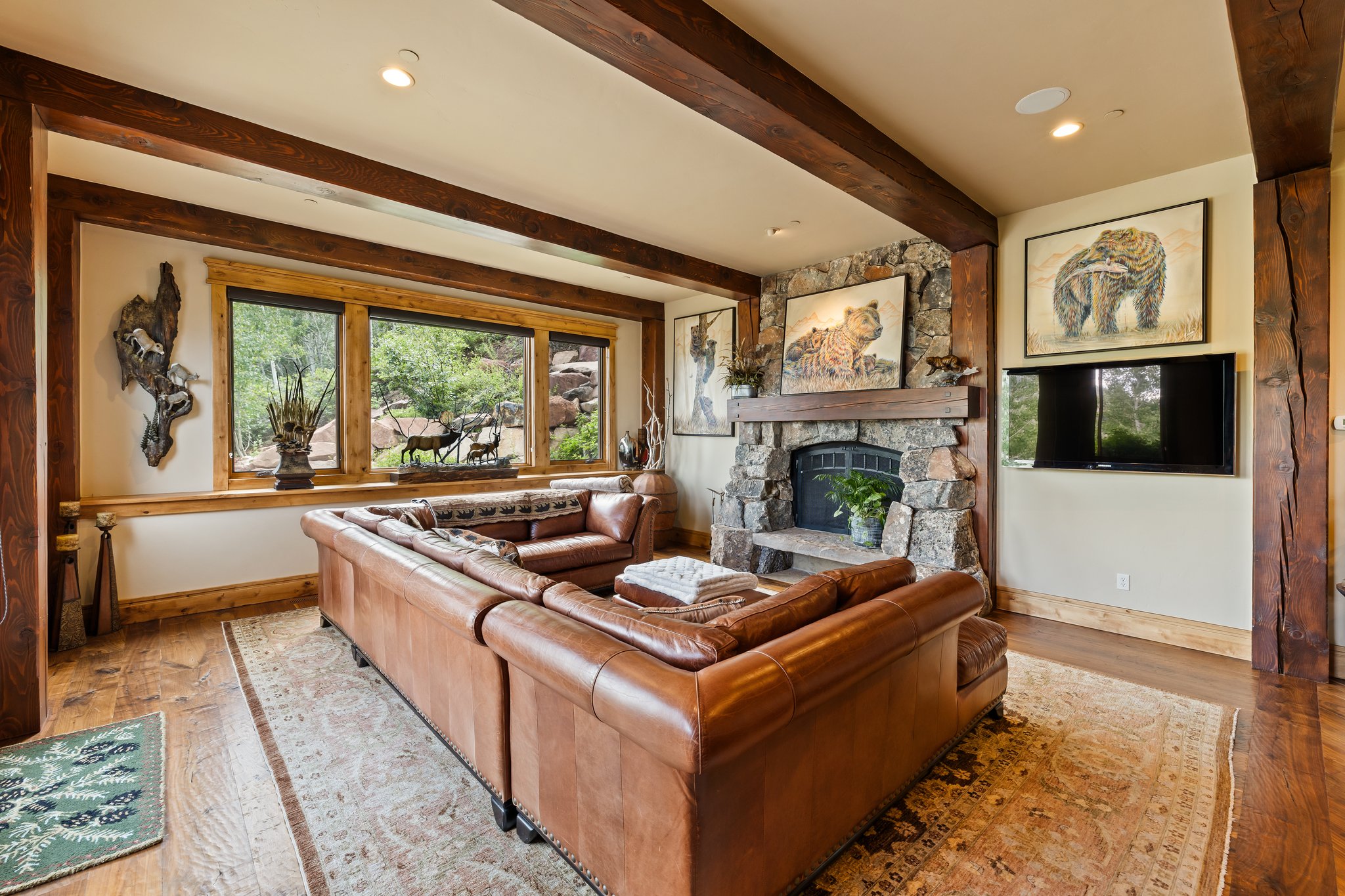 Spacious living area featuring exposed wood beams, a stone fireplace, wood-finish flooring, and a large window overlooking natural scenery