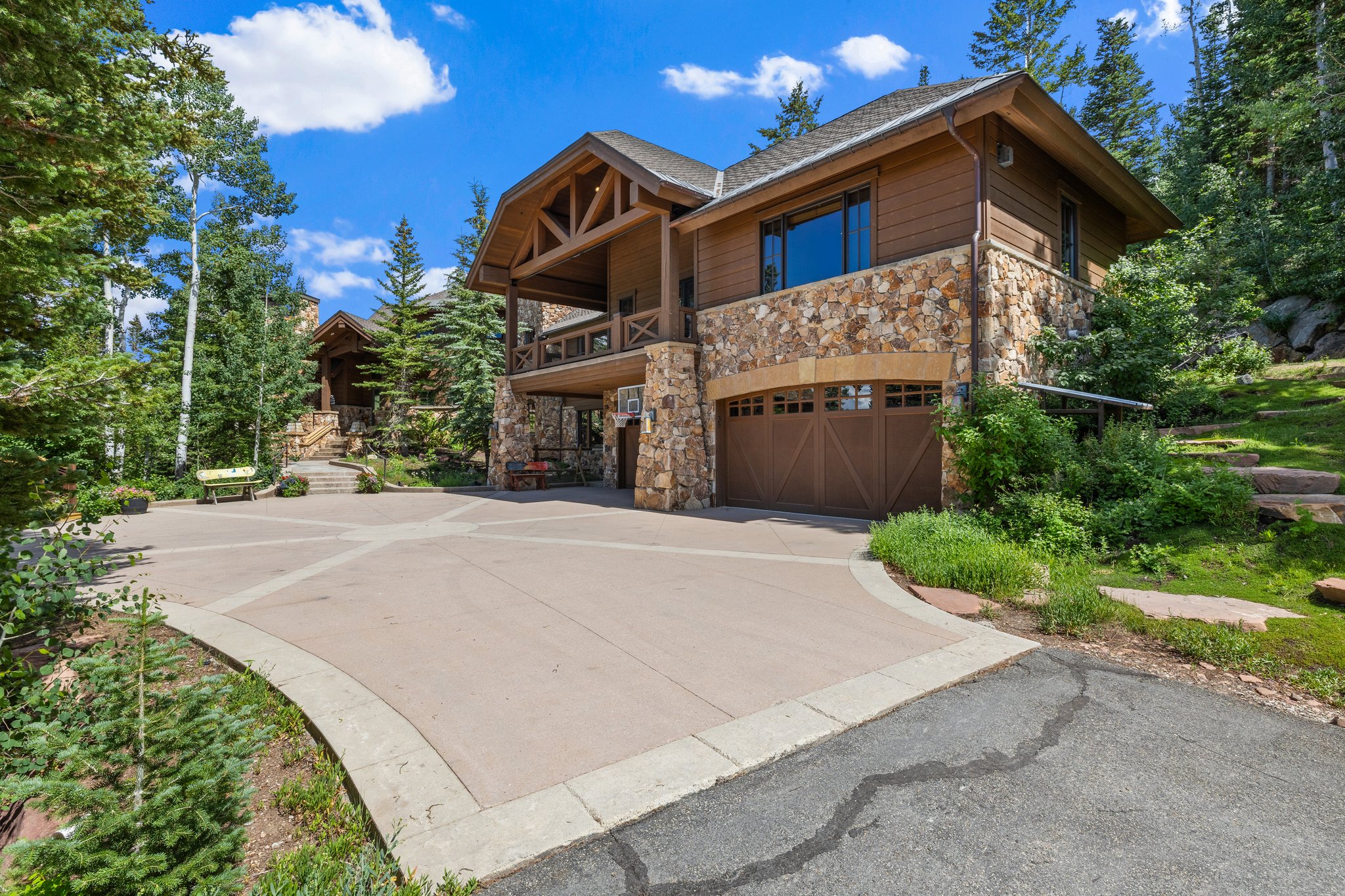 Stone and timber facade residence featuring a multi-level design, an attached garage with wood-finish doors, a prominent balcony, and a spacious paved driveway